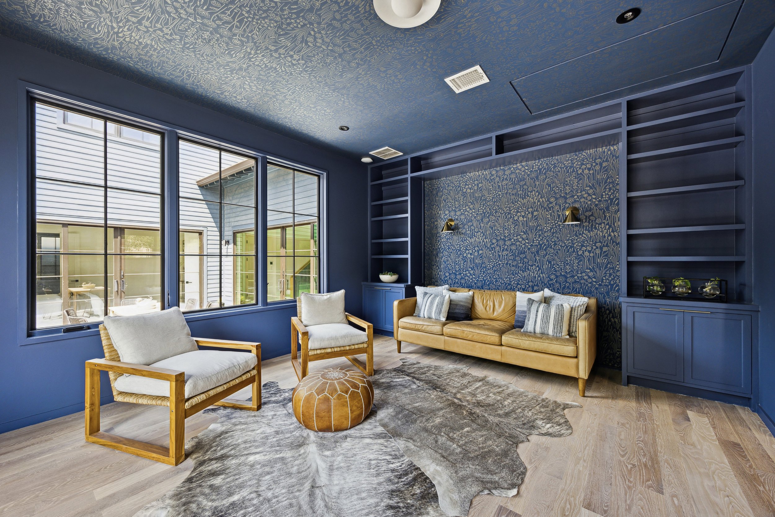 Living room with dark blue walls, large windows, light wood flooring, a yellow leather sofa with pillows, two armchairs with light cushions, a cowhide rug, built-in bookshelves, and modern wall lamps.