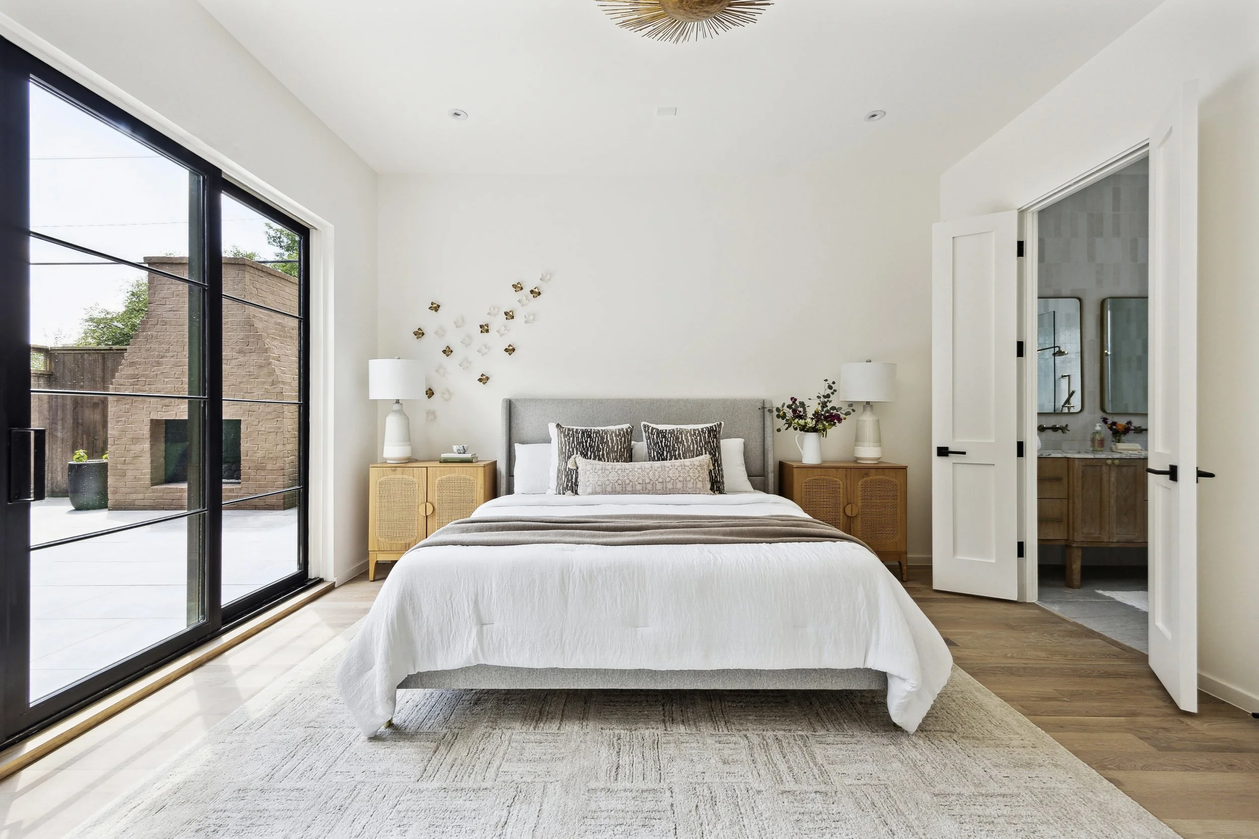 A modern bedroom with a large gray bed, white bedding, and decorative pillows. Two wooden bedside tables with white lamps and vases of flowers, large sliding glass door to an outdoor patio, and a doorway leading to a bathroom.