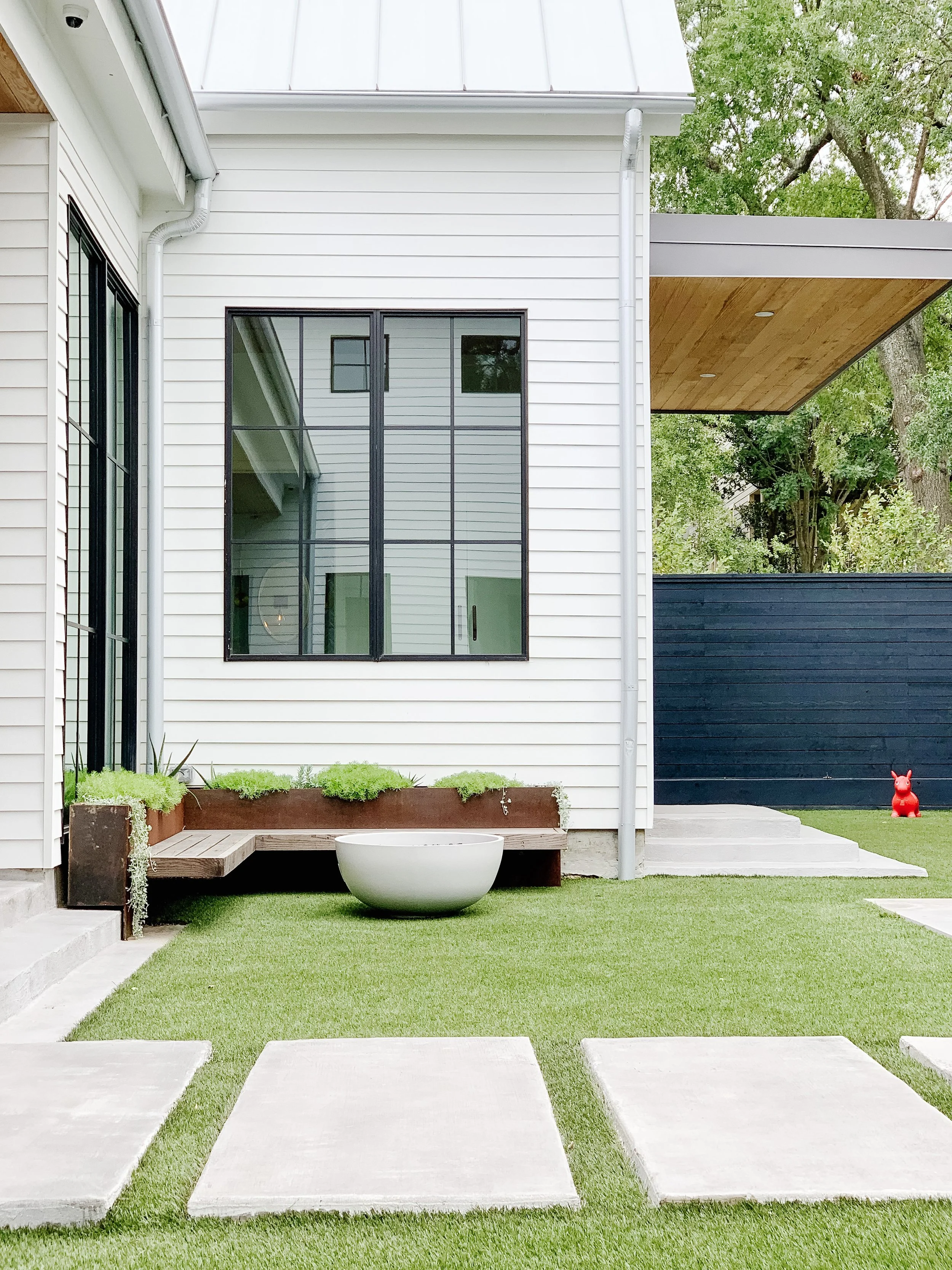 Modern backyard with concrete stepping stones on green grass, planter with greenery, large window, wooden bench, white and black exterior walls, red dog statue, and trees in the background.
