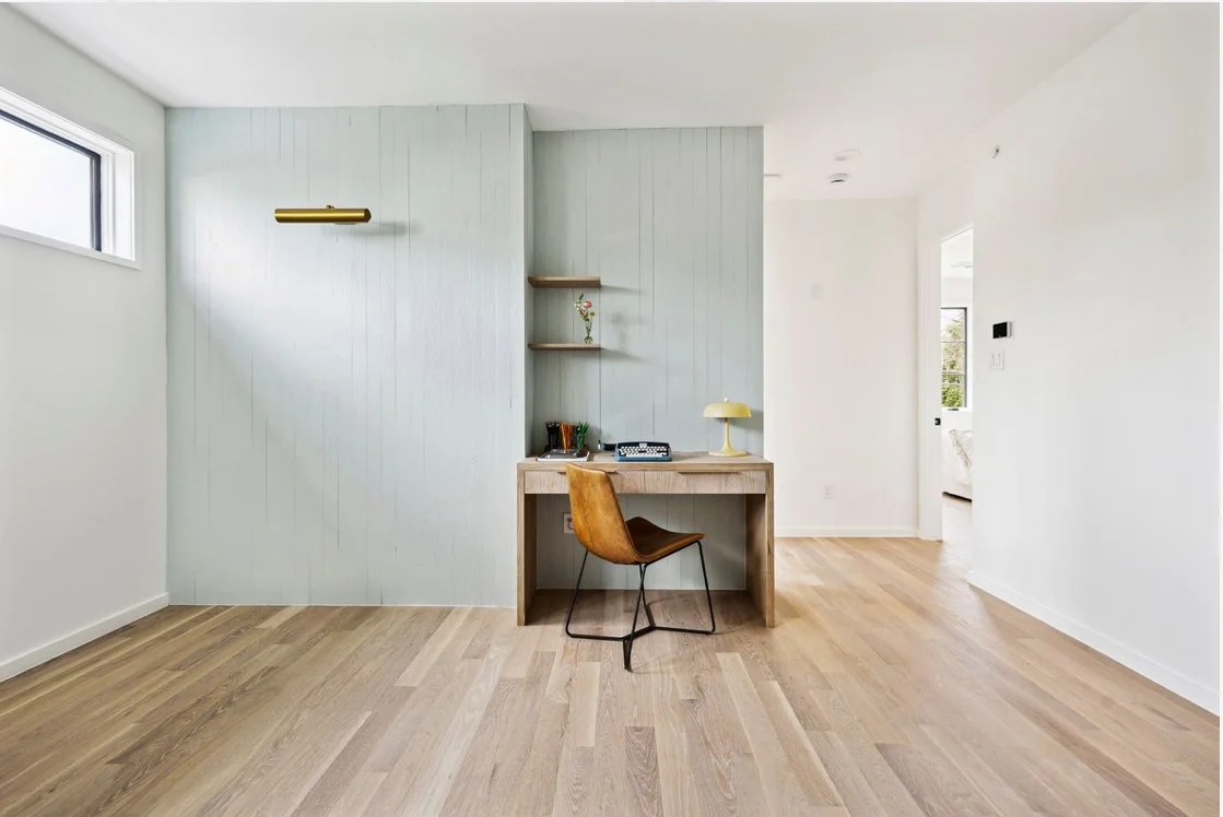 Minimalist home office with a small wooden desk, a leather chair, a yellow desk lamp, and floating shelves with decorative items, in a bright, airy room with white walls and light wood flooring.