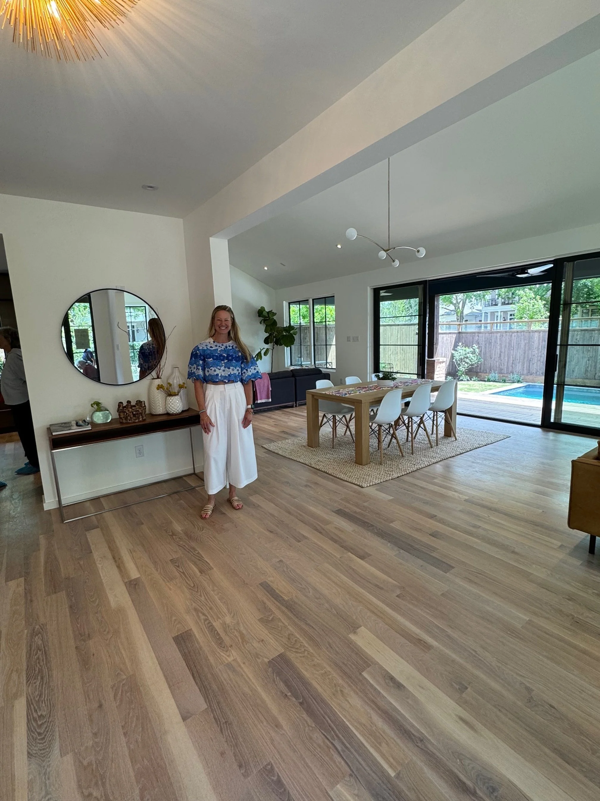 A woman standing in a modern, spacious living and dining area with wooden flooring, a dining table with white chairs, a black sofa, large windows, and sliding glass doors leading to an outdoor pool.