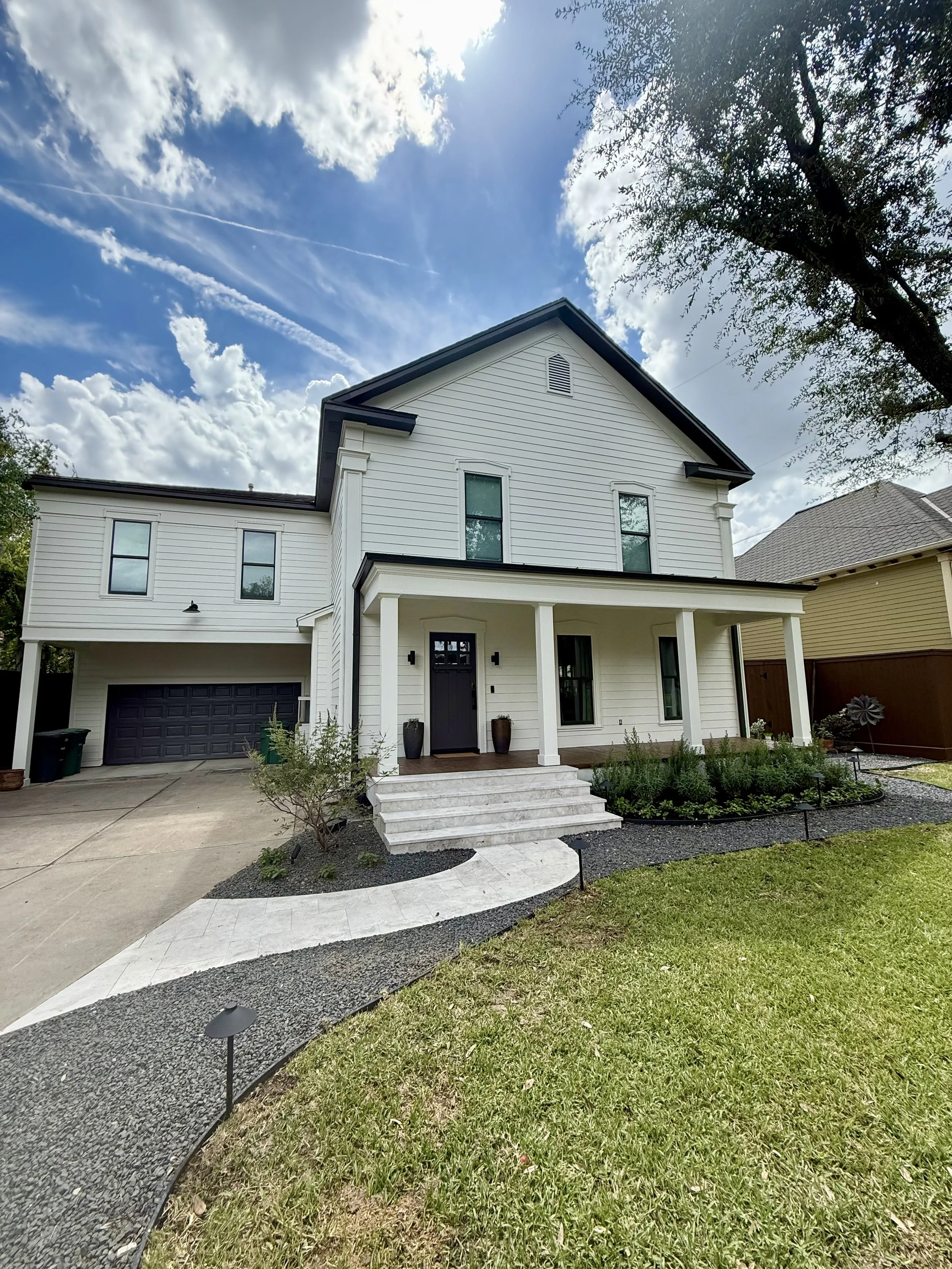 White two-story house with front porch, black door, and attached garage, surrounded by a manicured lawn, small bushes, and landscaping lighting under a partly cloudy sky.