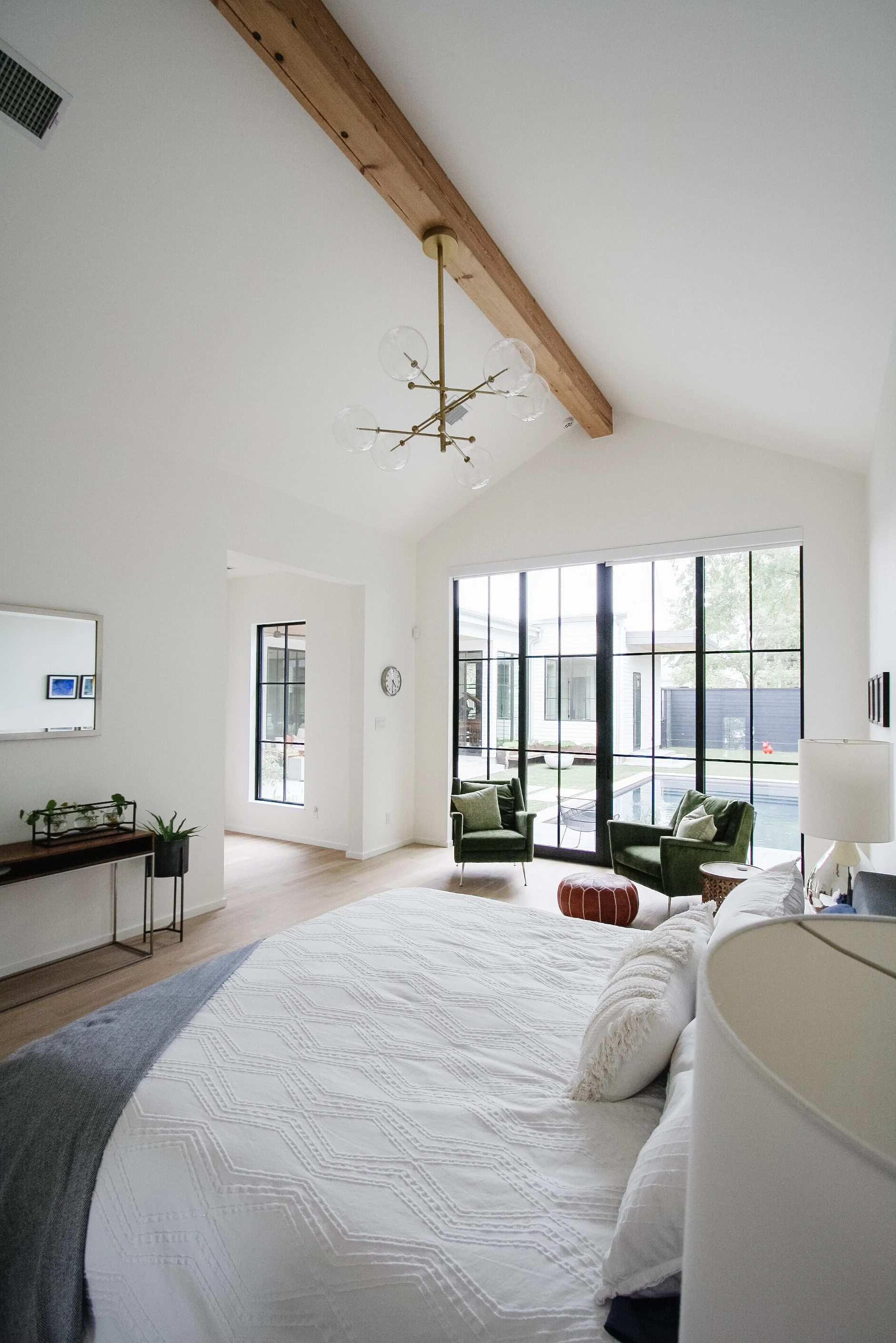 Bright bedroom with white walls, a large bed with white bedding, a wooden bench, a mirror, two green armchairs by large glass windows, and a view of a pool outside.