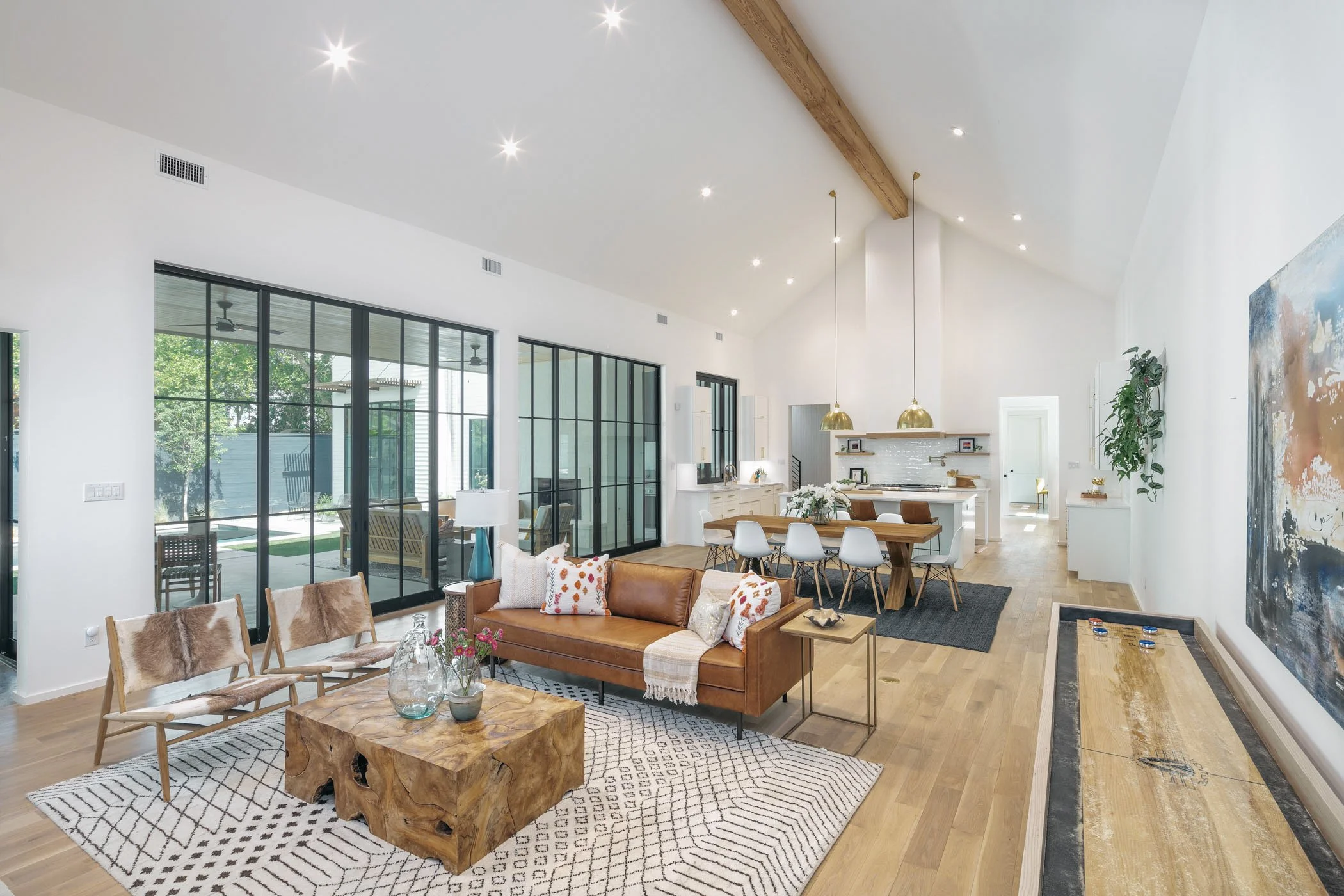 Spacious open-concept living and dining area with large windows, white walls, wooden beams, and modern furniture, including a brown leather sofa, wooden chairs with gray cushions, and a farmhouse table with white chairs. Decor includes colorful pillows, a patterned rug, a wooden coffee table with vases, and abstract wall art.