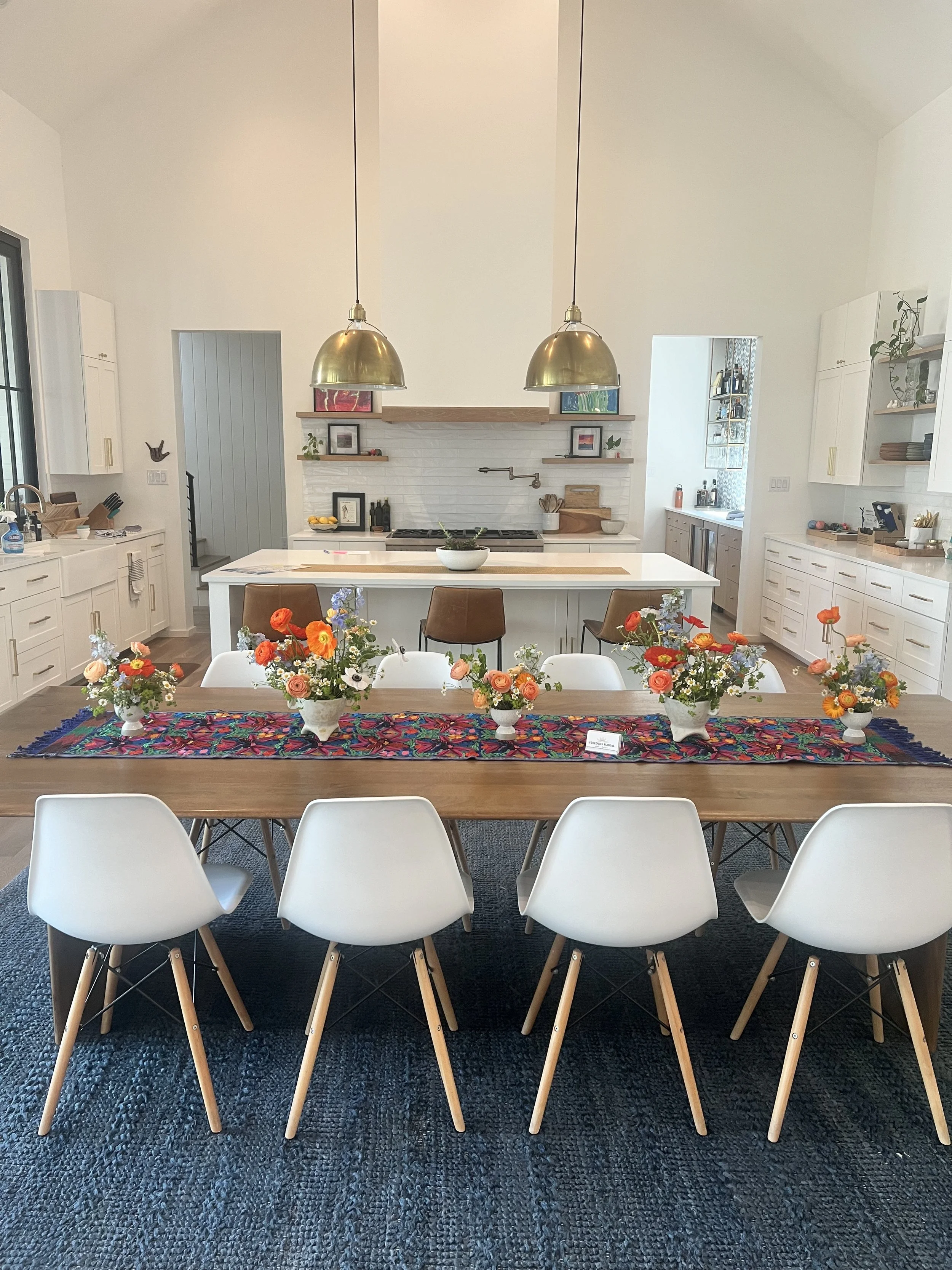 A dining room featuring a wooden table with a colorful floral table runner and five white chairs, with five small flower arrangements. The kitchen in the background has white cabinets, a brown island with a white countertop, and two large brass penda