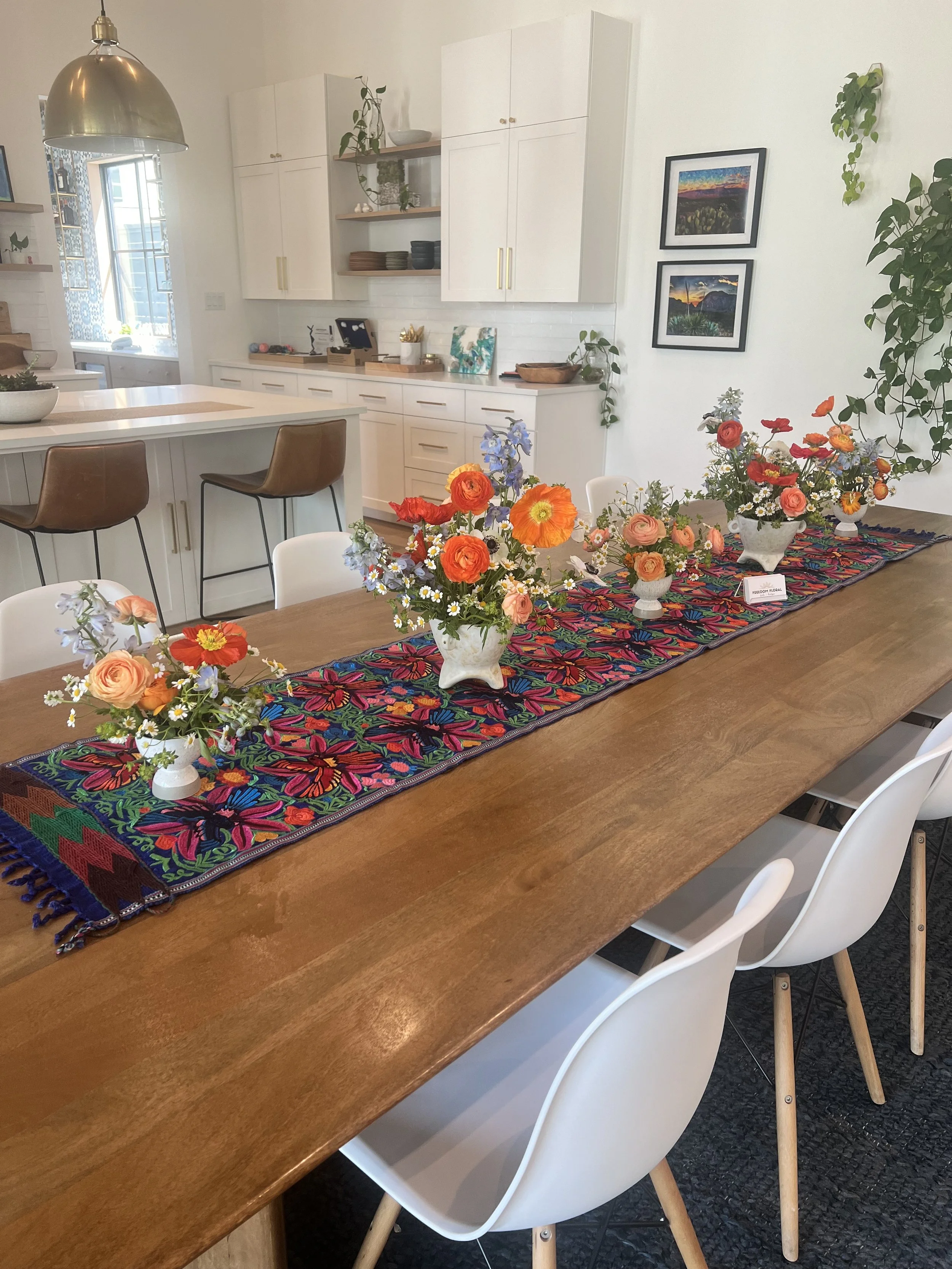 A dining table decorated with colorful floral arrangements in white vases, placed on an embroidered table runner, with a modern kitchen in the background.