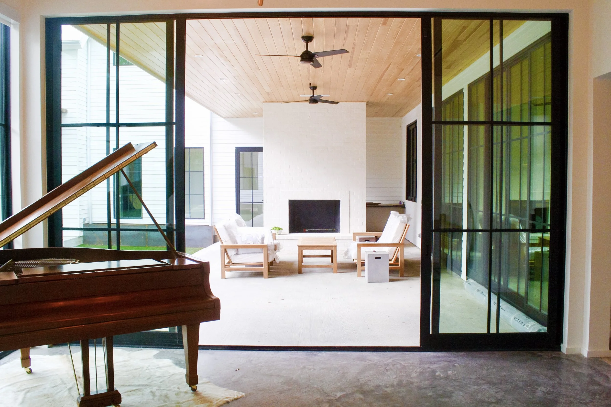 Living room with a white brick fireplace, wooden chairs with white cushions, and a white sofa. Large sliding glass doors open to a patio. There are ceiling fans and a rustic wooden ceiling.