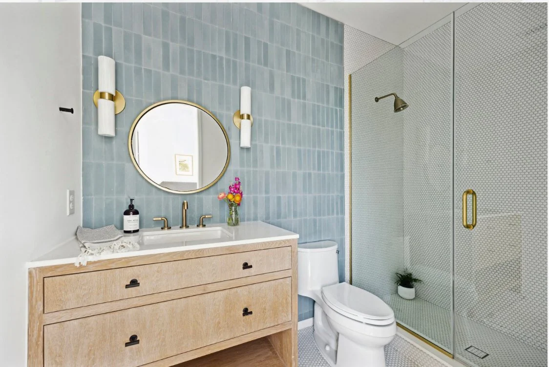 Modern bathroom with a wooden vanity, round mirror, blue tiled wall, and glass shower enclosure with gold accents, potted plant, and colorful flowers.