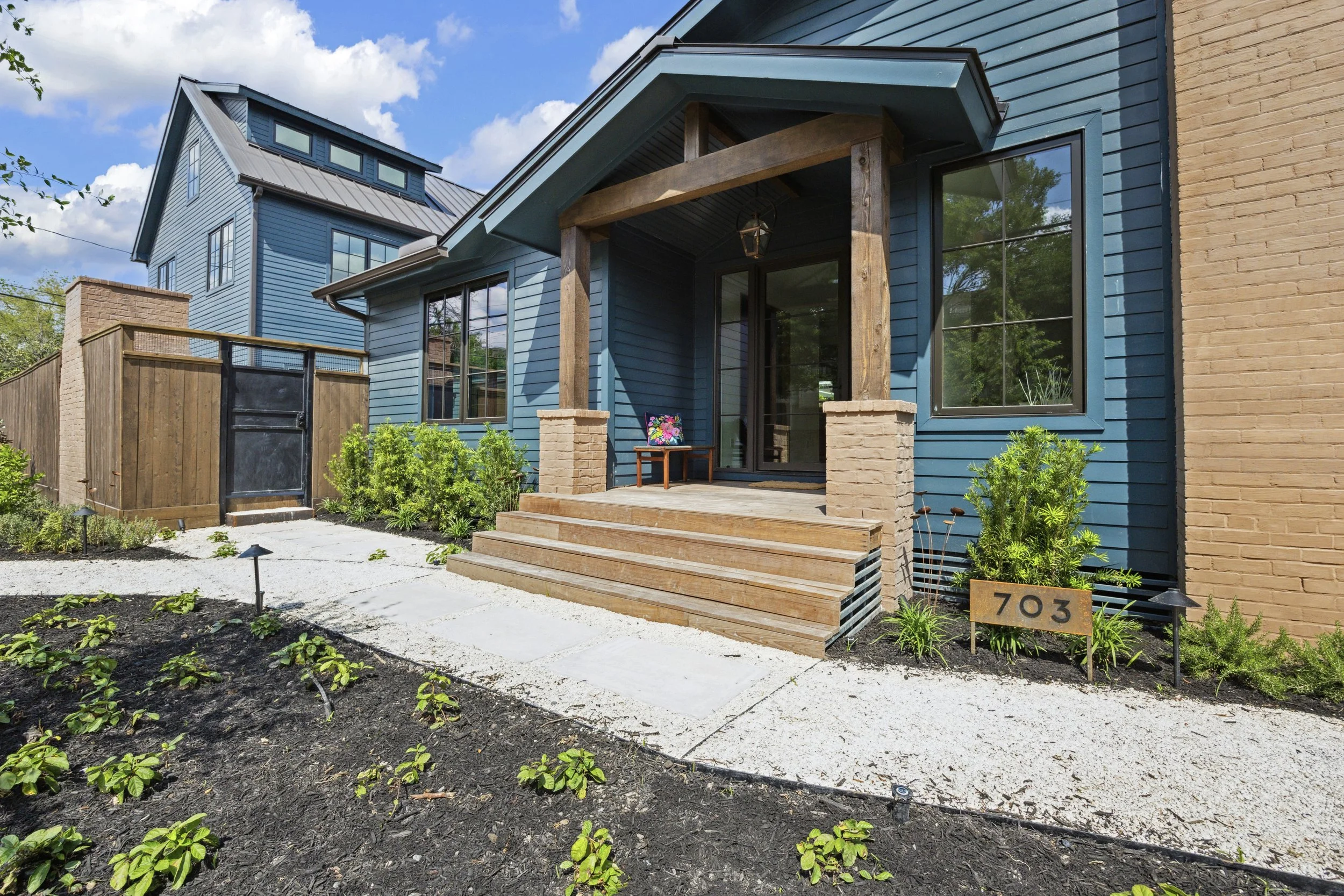 A modern two-story house with blue and beige exterior walls, large windows, a small porch with wooden steps, and landscaped garden with small plants and bushes.