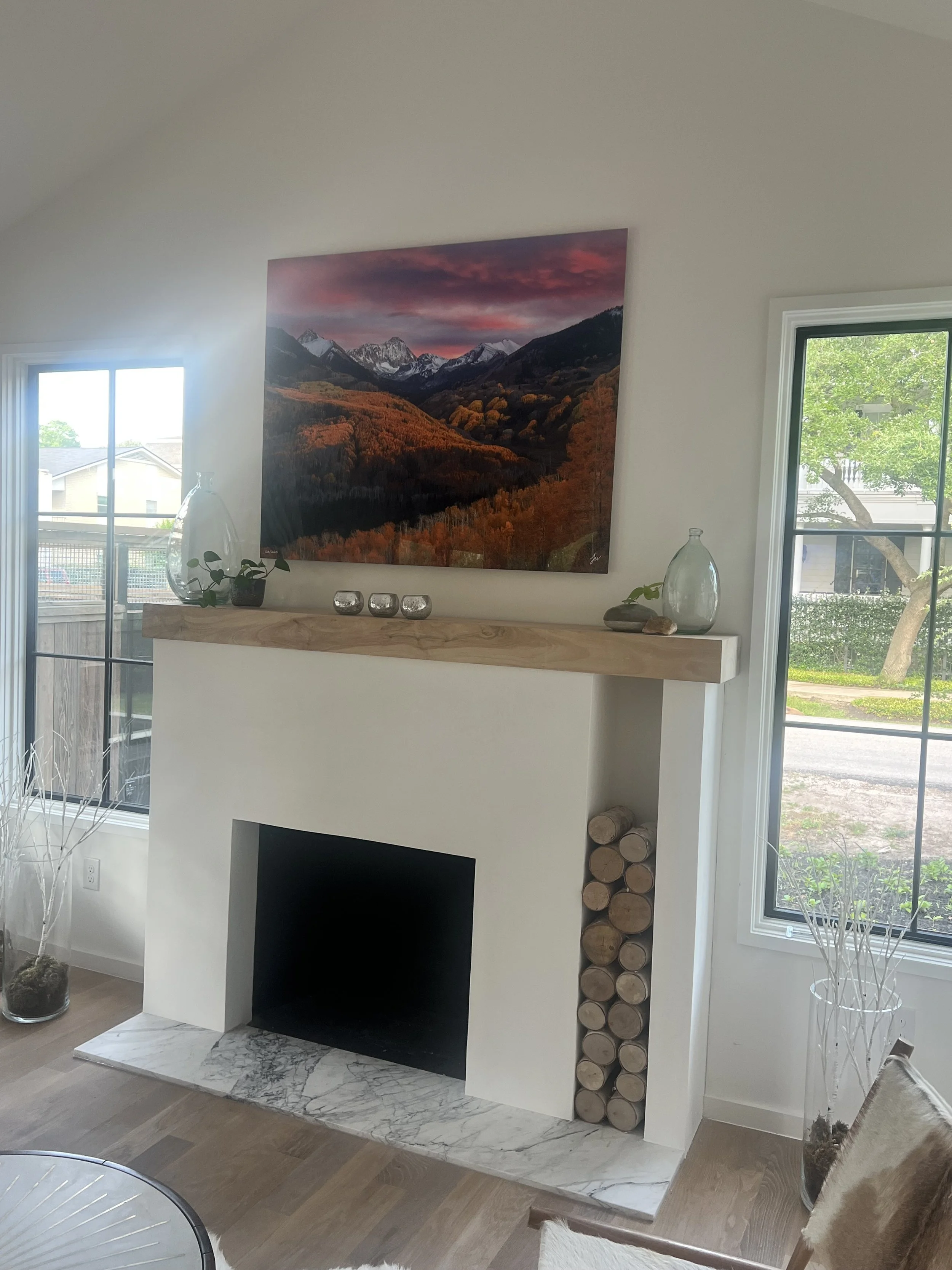 Living room with a white fireplace, a wooden mantel decorated with glass vases and candles, logs stored on the side, a landscape painting, and windows showing trees and neighborhood outside.