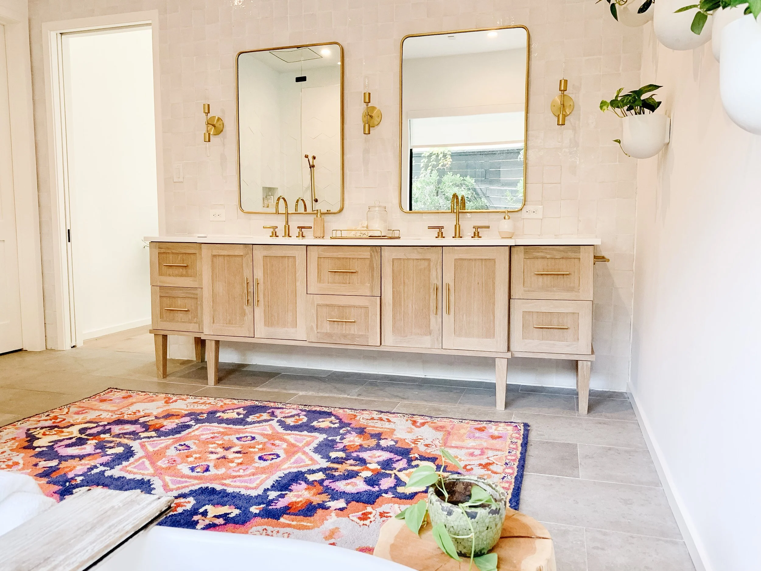A modern bathroom vanity with two mirrors, gold fixtures, and light wood cabinetry. There are wall-mounted planters with green plants on the right, and a colorful rug on the floor.