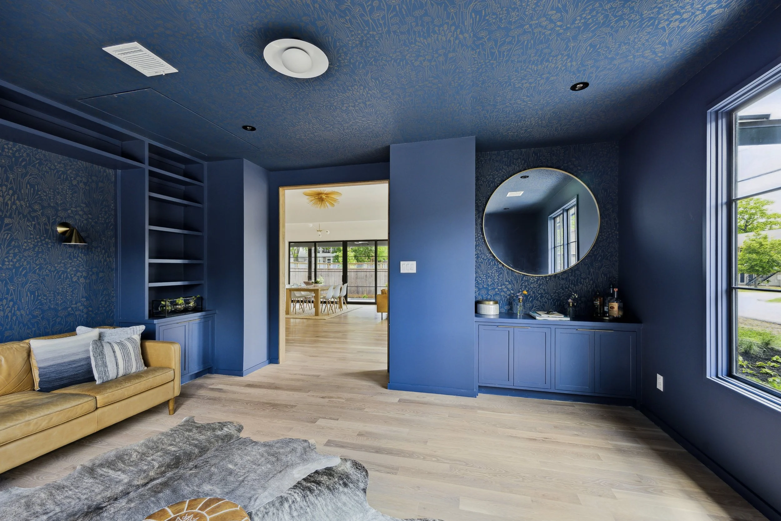 Modern living room with navy blue walls, light wood flooring, a large window, a circular mirror, a tan leather couch with striped pillows, and a cowhide rug.
