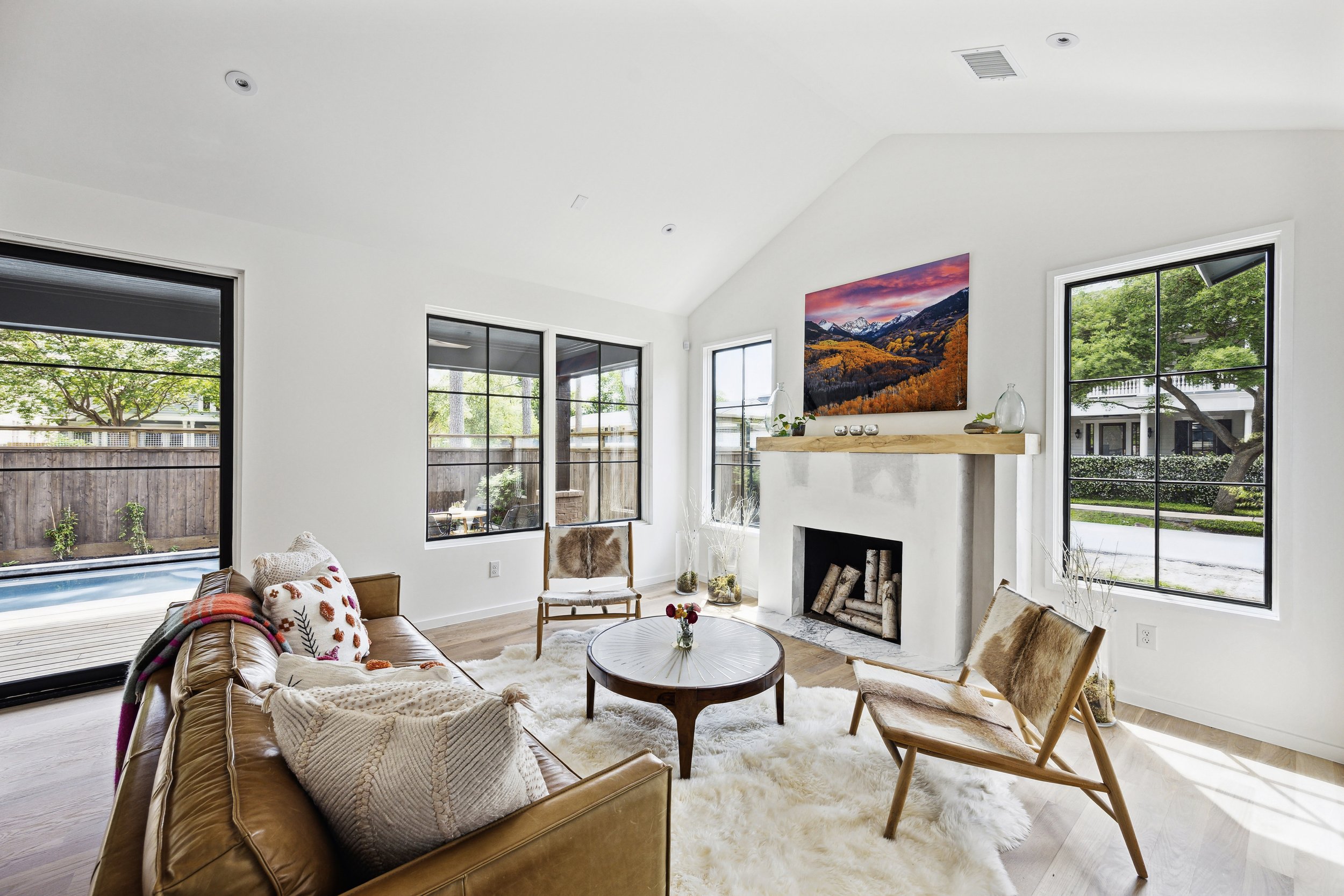 Bright living room with white walls, large windows, a tan sofa with patterned pillows, a white fluffy rug, wooden chairs with fur cushions, a round coffee table, and a fireplace with a mounted TV displaying a mountain landscape.