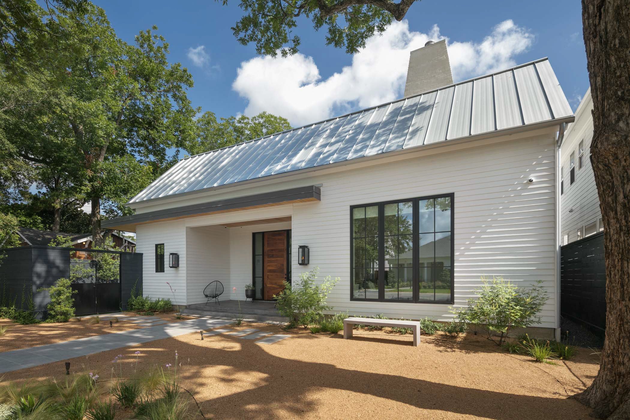 Modern white house with black window frames and a metal roof, surrounded by trees, with a sandy front yard, a black fence on the left, and a wooden door, under a partly cloudy sky.