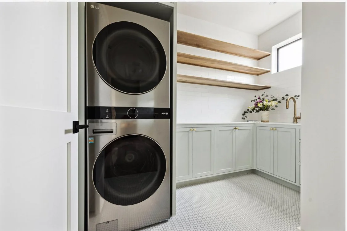 Laundry room with stacked washer and dryer, white cabinets, open wooden shelves, a small window, and a countertop with a vase of flowers