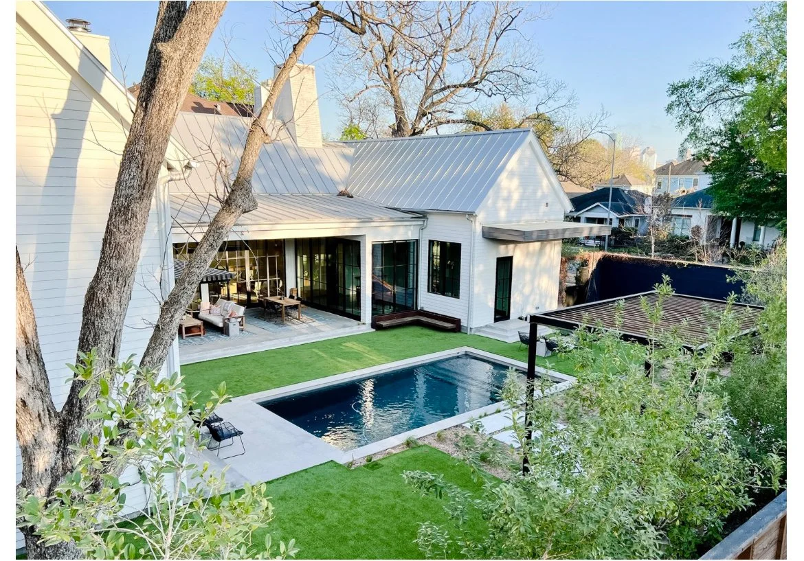 A modern white house with a metal roof, featuring a backyard with a rectangular swimming pool, a lush green lawn, patio furniture, and trees surrounding the yard.