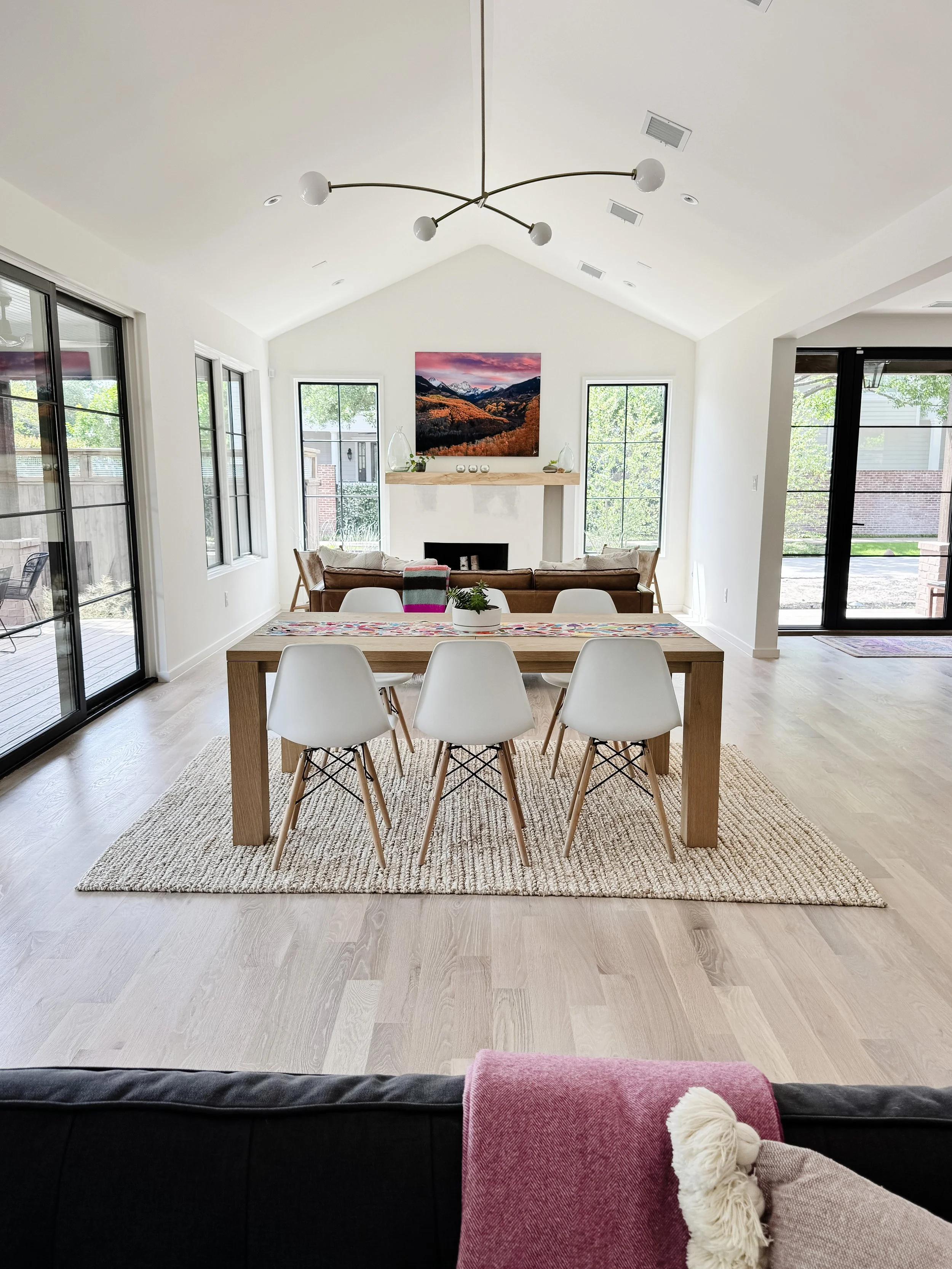 Bright open-concept living and dining room with white walls, light wood flooring, and large windows. A wooden dining table with white chairs sits on a rug. The living area features a fireplace with a forest landscape painting above, and a couch with pillows and a throw blanket. Sliding glass doors lead to an outdoor patio.