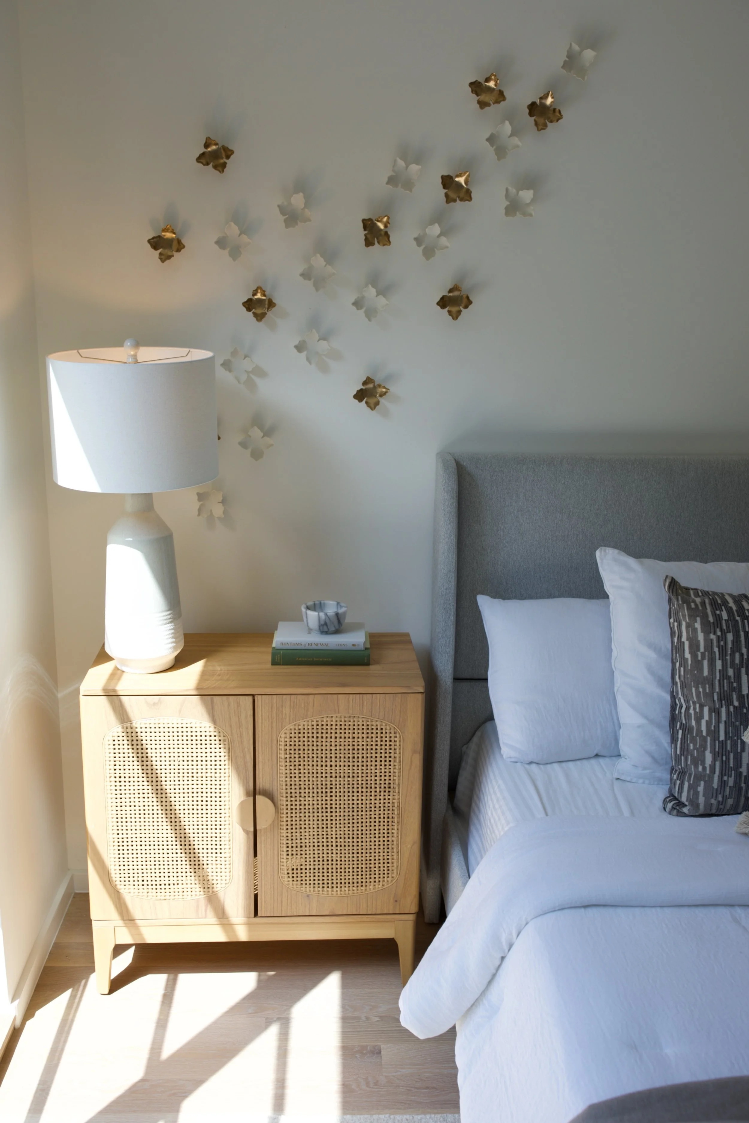 Bedroom corner with a wooden nightstand, a white table lamp, a bowl, and books, next to a gray upholstered bed with pillows, and decorative metallic flower wall art.