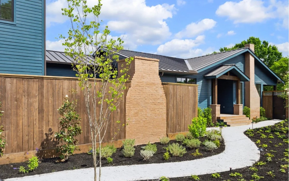Exterior of a modern house with blue siding, brown brick chimney, and a landscaped yard with a curved white gravel pathway, young trees, and small bushes under a partly cloudy sky.