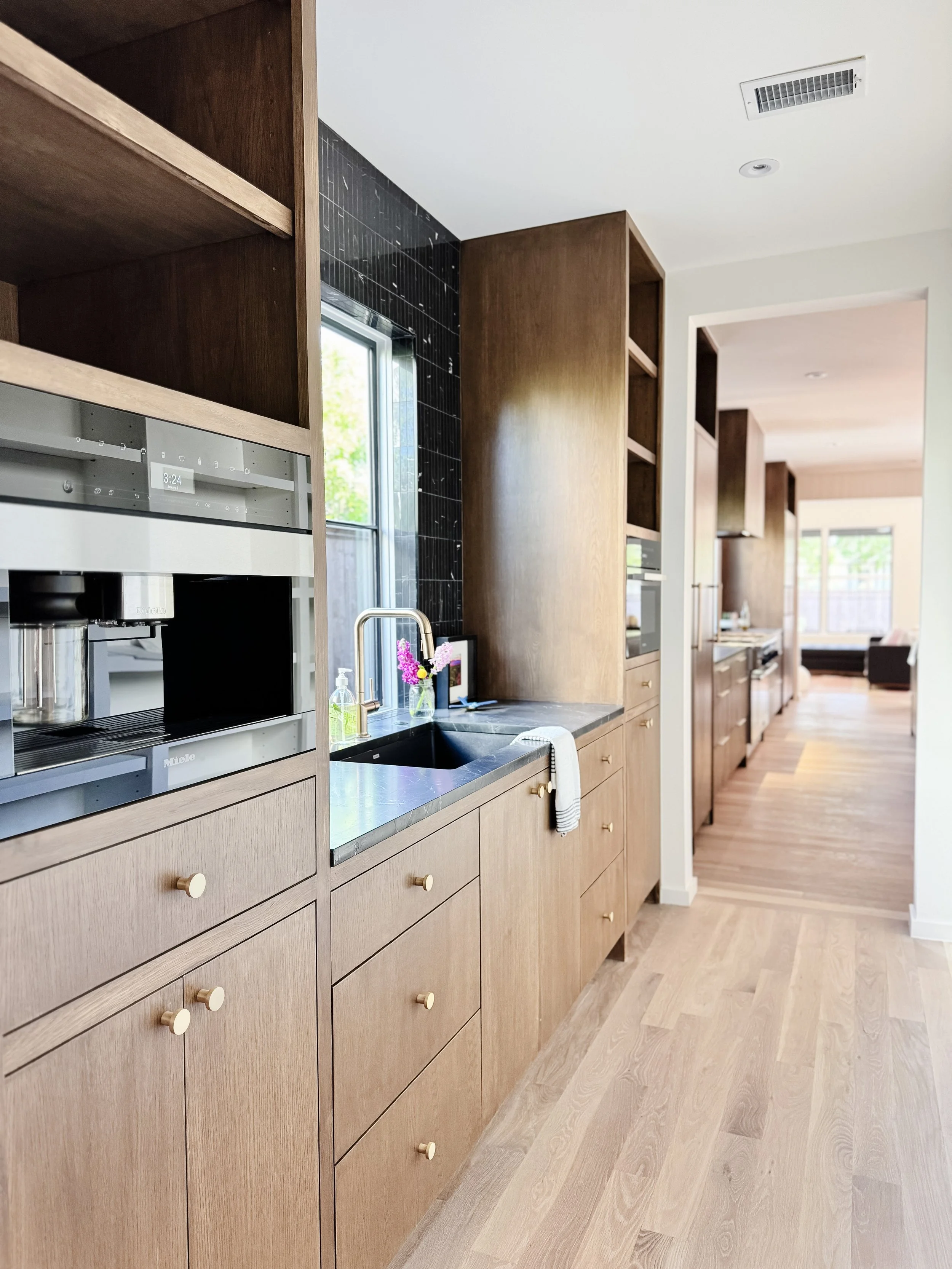 Modern kitchen with wooden cabinets, a black countertop, and a stainless steel coffee machine. A small vase with pink flowers is near the sink, and a white dish towel hangs on a cabinet handle. An opening shows a bright living area with large windows and hardwood floors.