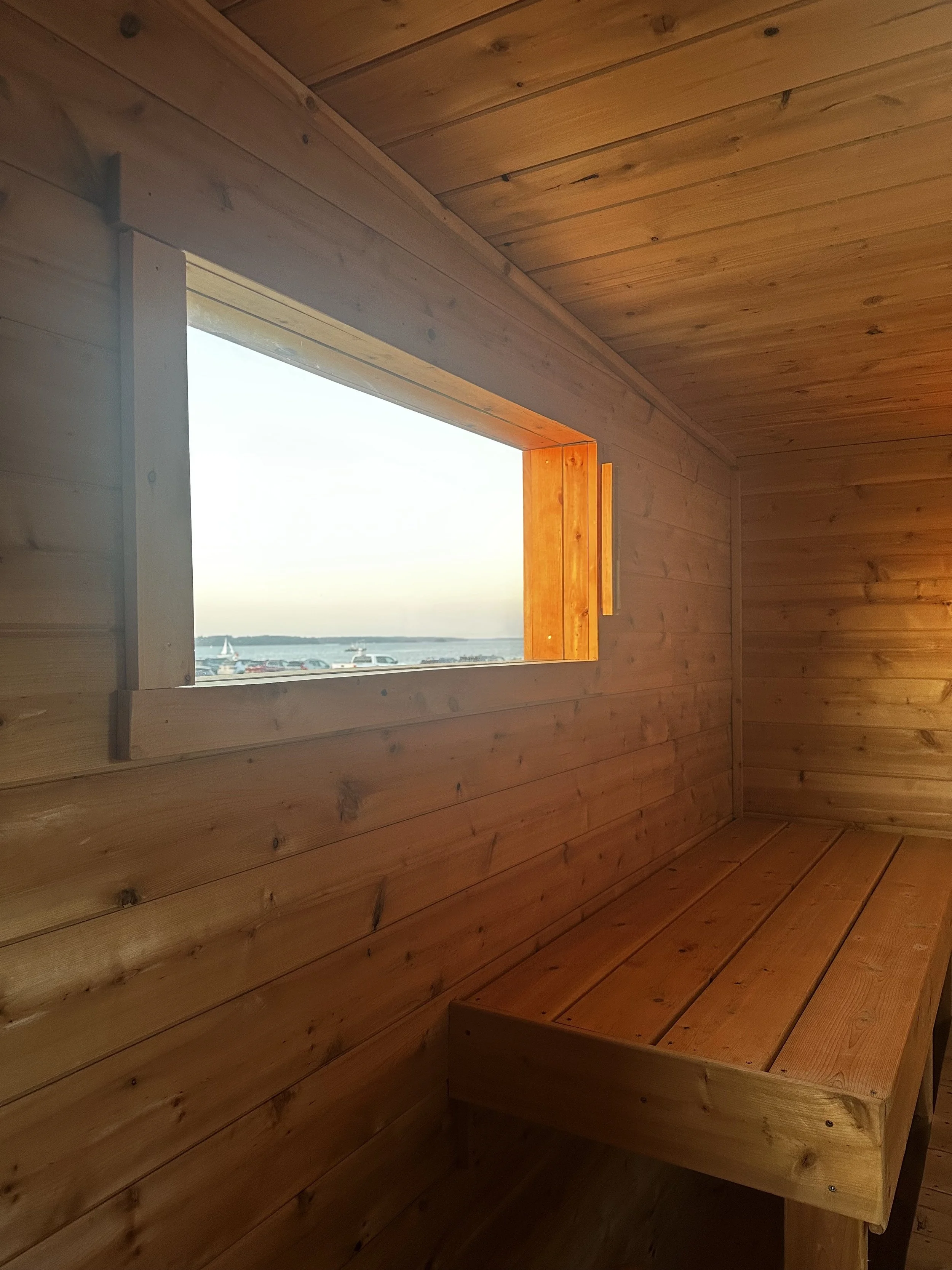 Inside a wooden structure with a rectangular window revealing a view of boats and water outside, featuring wooden walls and ceiling with a built-in wooden bench.