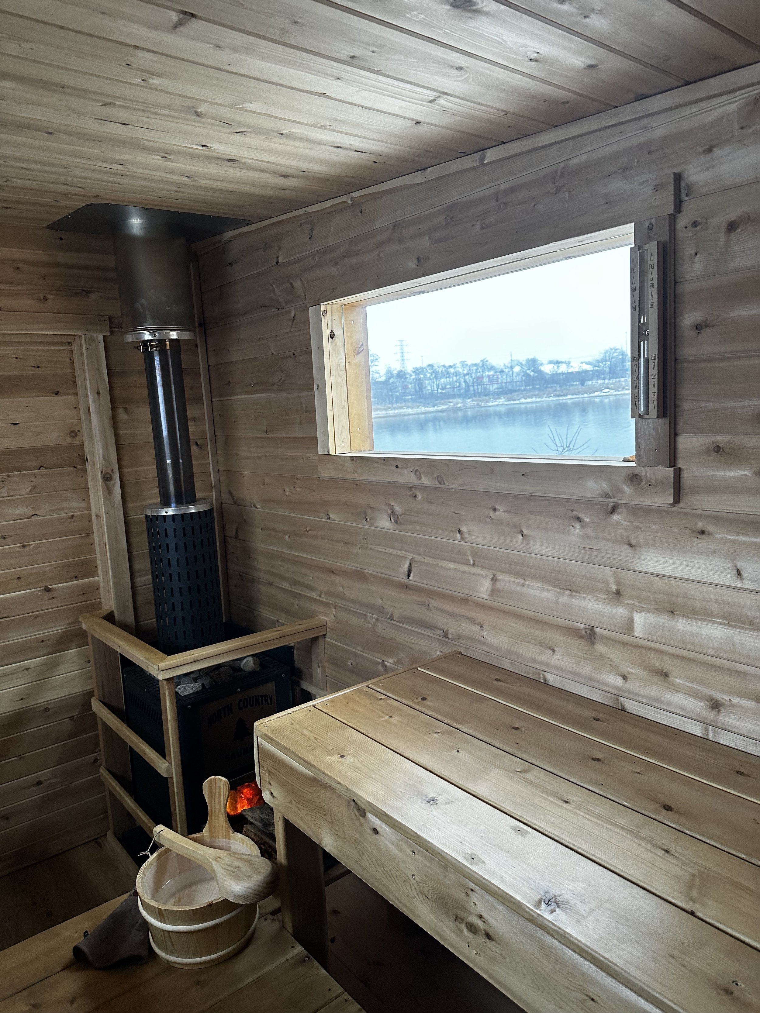 Interior of a small wooden sauna with a wooden bucket and ladle, a bench, a stove, and a rectangular window overlooking a lake and trees.