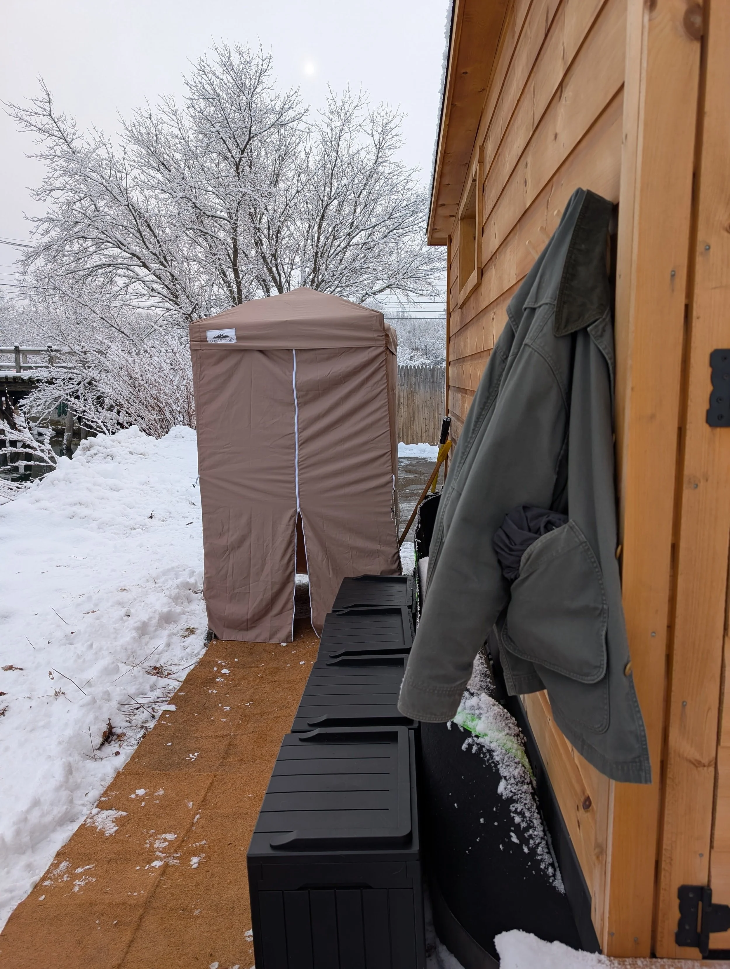Snow-covered backyard with a wooden shed, a brown outdoor storage tent, and a black storage bench. A gray jacket hangs on the shed's exterior. Snow surrounds the area, and a large, leafless tree is in the background under an overcast sky with the sun visible.