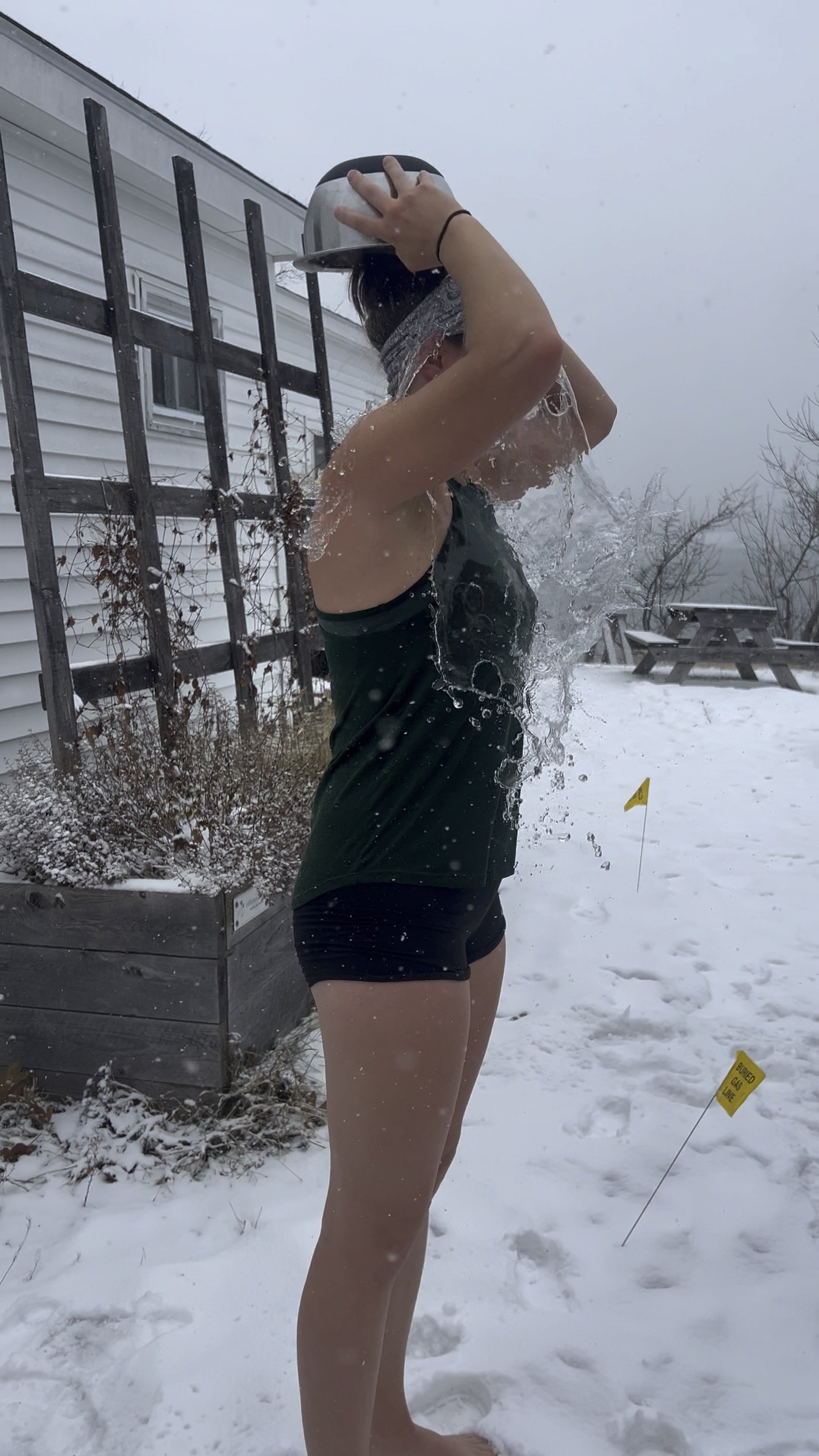 A person standing barefoot in the snow, rinsing their hair with water from a gray bowl, with snow-covered ground and outdoor furniture in the background.