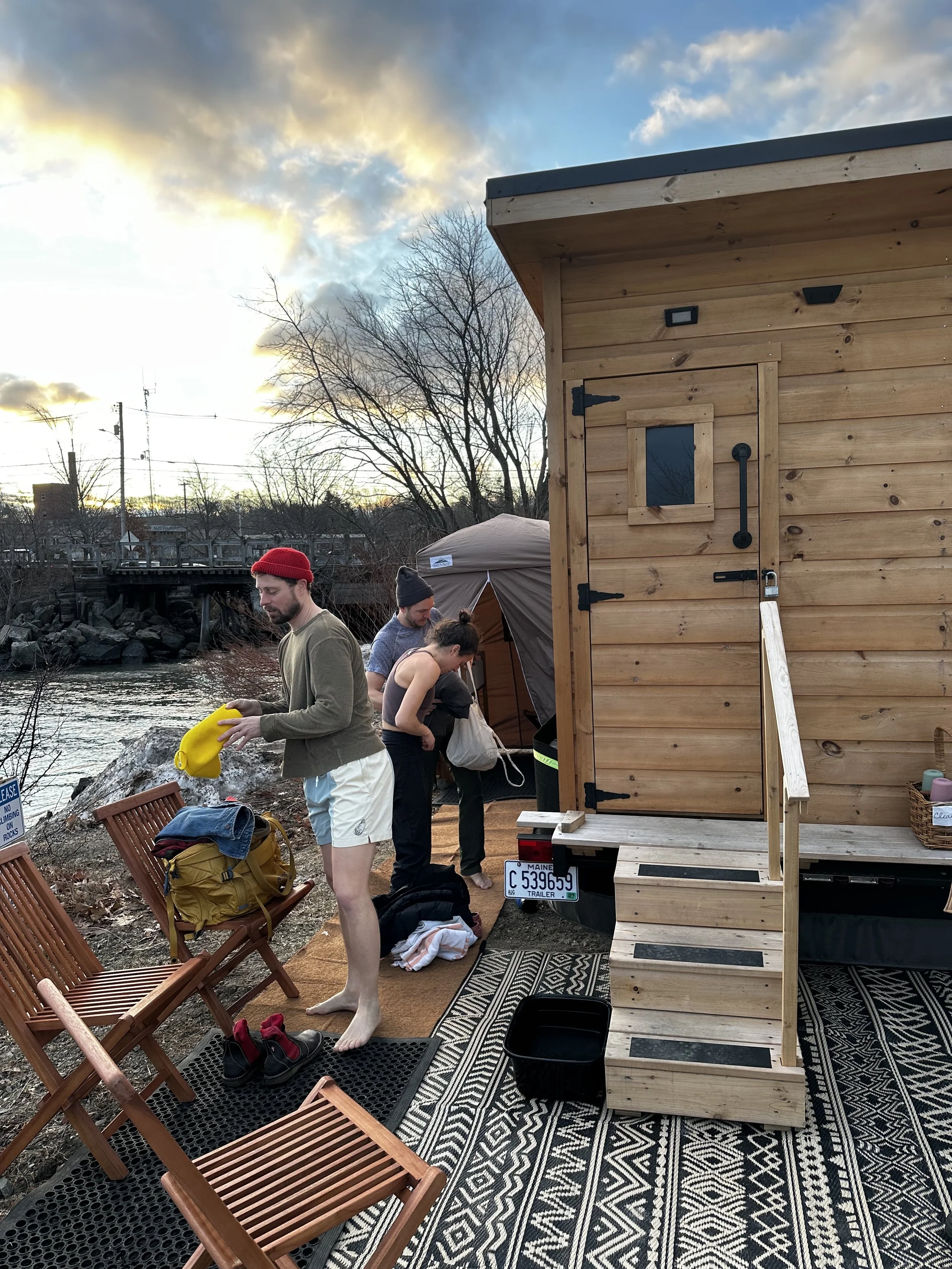 People on a dock near a river, with a small wooden house, trees, and a cloudy sky in the background. One person is watering with a yellow watering can, and others are preparing or organizing gear.