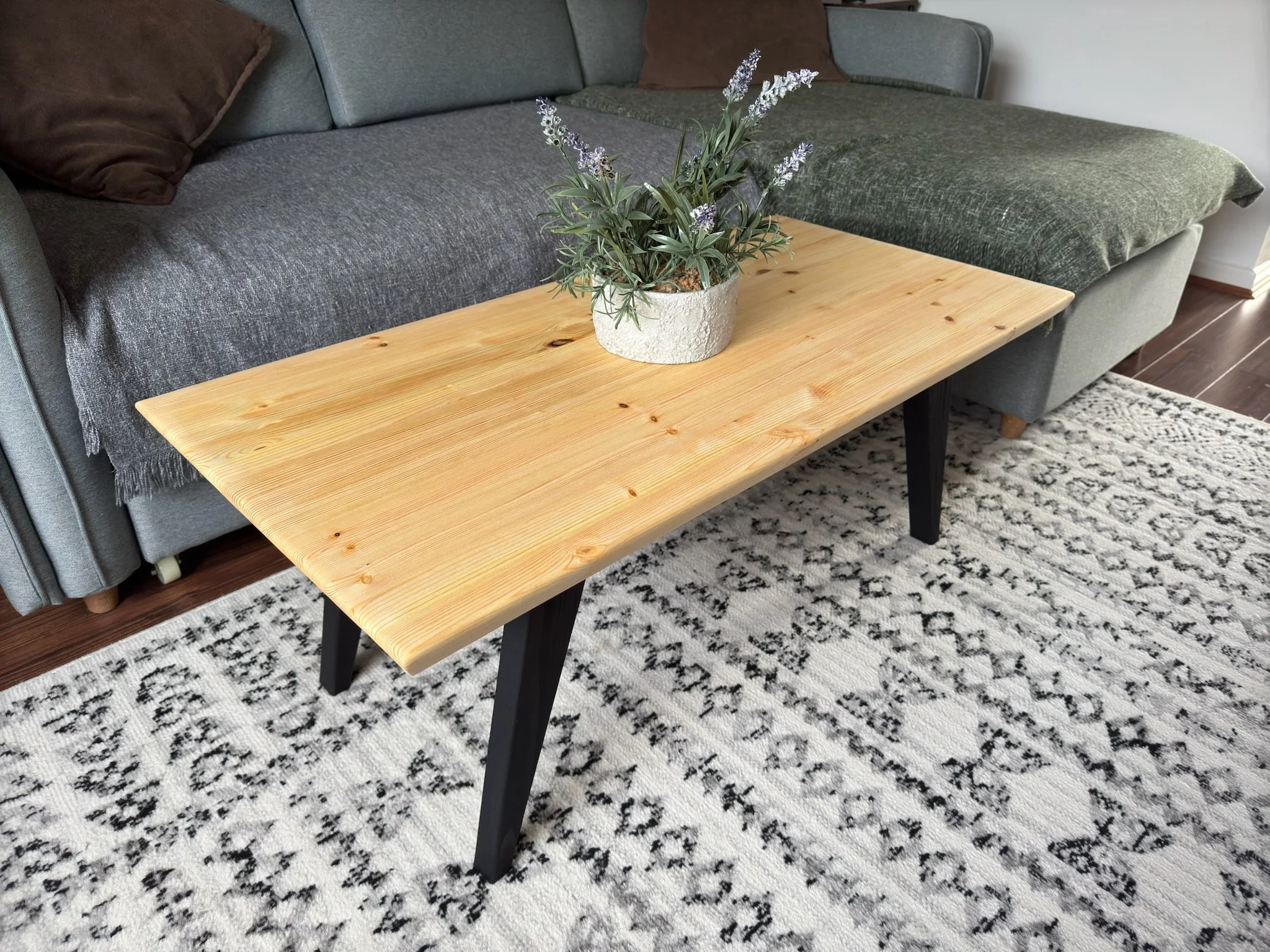 A wooden coffee table with black angled legs, placed on a patterned black and white rug in front of a green sofa with brown pillows, and a potted plant with purple flowers on top.
