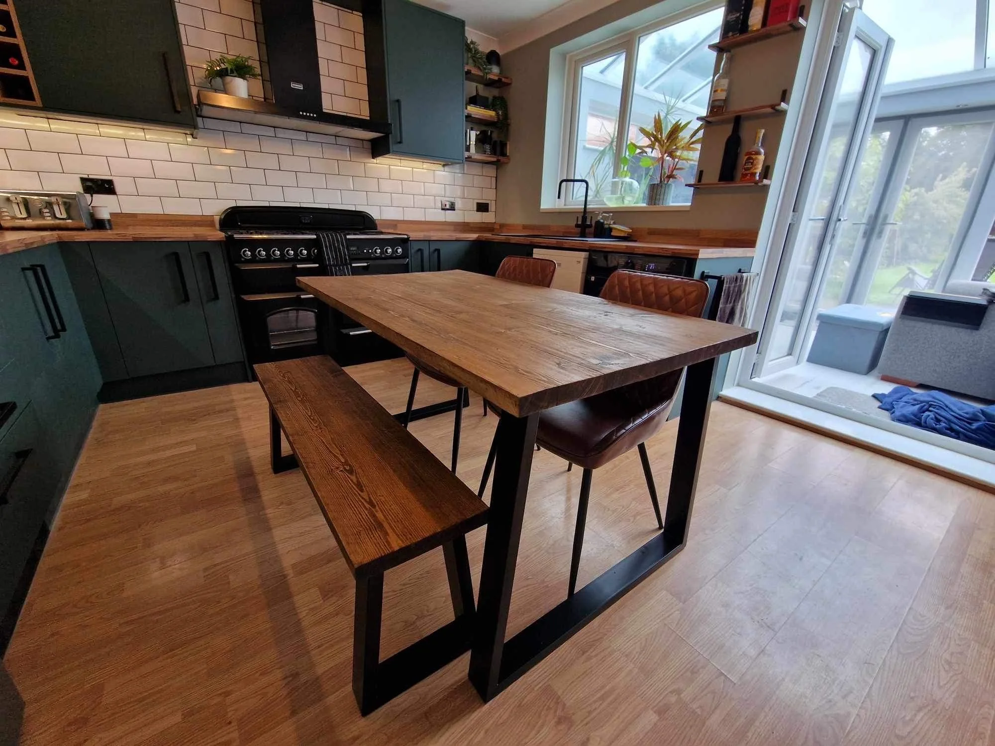Modern kitchen with dark green cabinets, wooden countertops, white subway tile backsplash, and natural light from large windows and glass door. Wooden dining table with a bench and chairs, plants, and decorative items.