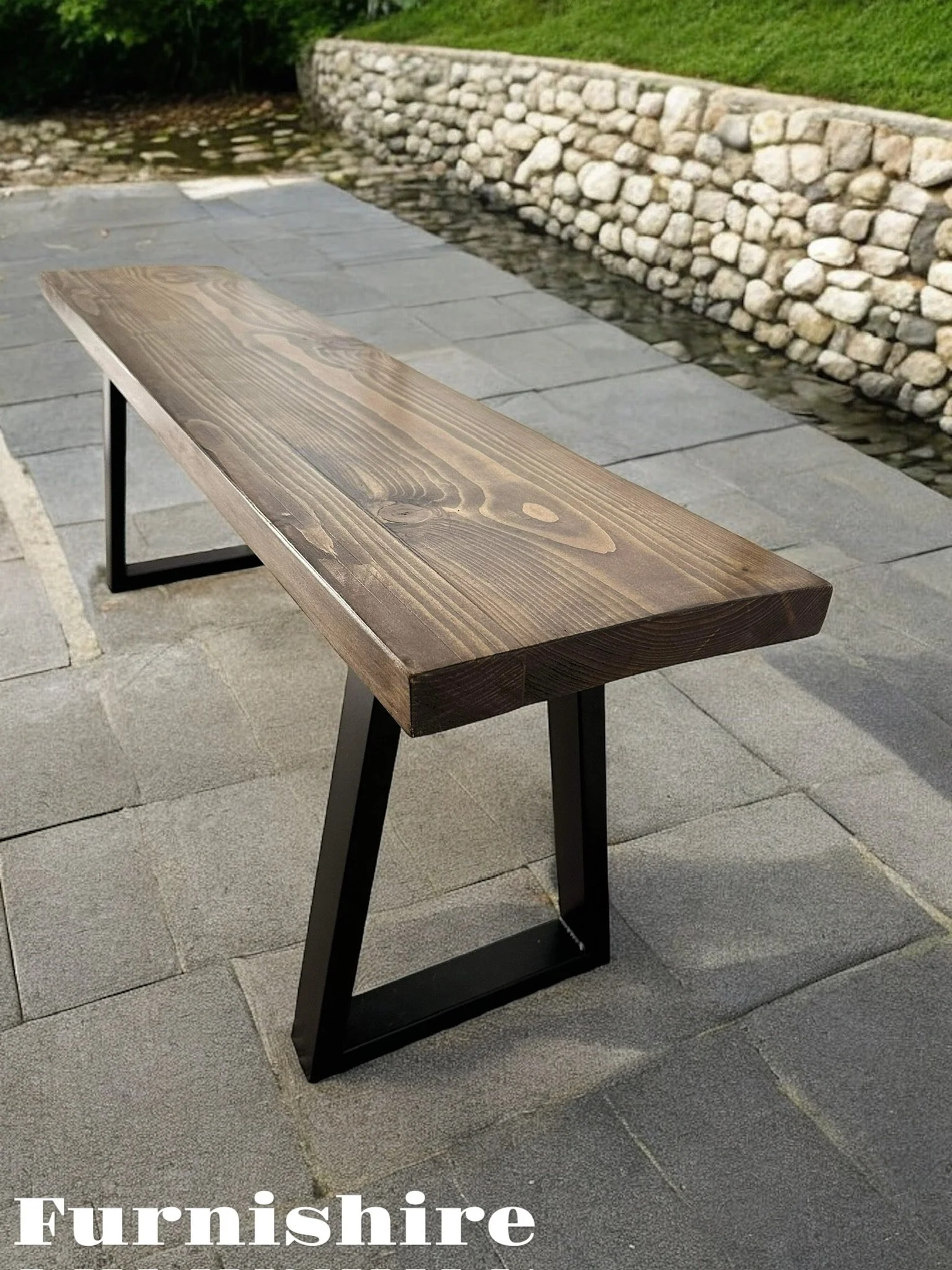 A wooden table with black metal legs outdoors on a stone-paved surface, with a stone wall and grassy area in the background.