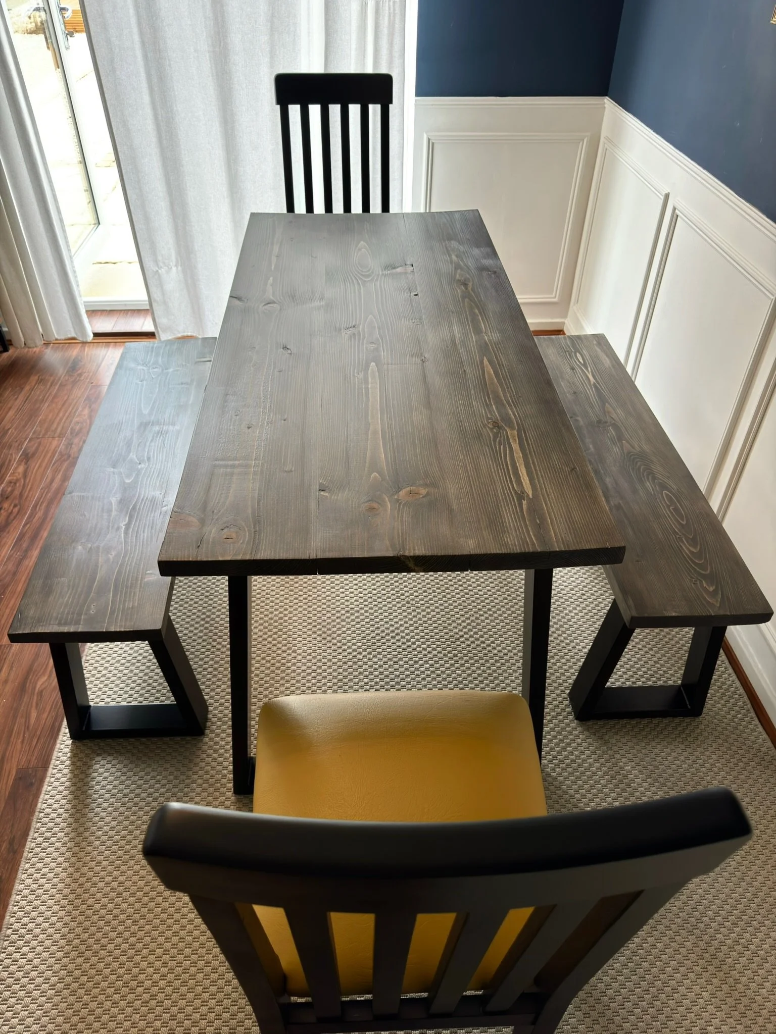 A dining table with a dark wood top, an open space with a sliding glass door, a chair with a yellow cushion, and a corner wall with white wainscoting and dark blue paint above.