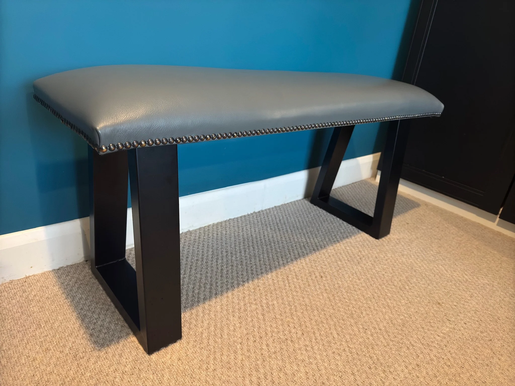 A modern beige upholstered bench with black wooden legs and nailhead trim, placed against a teal wall and beige carpeted floor.