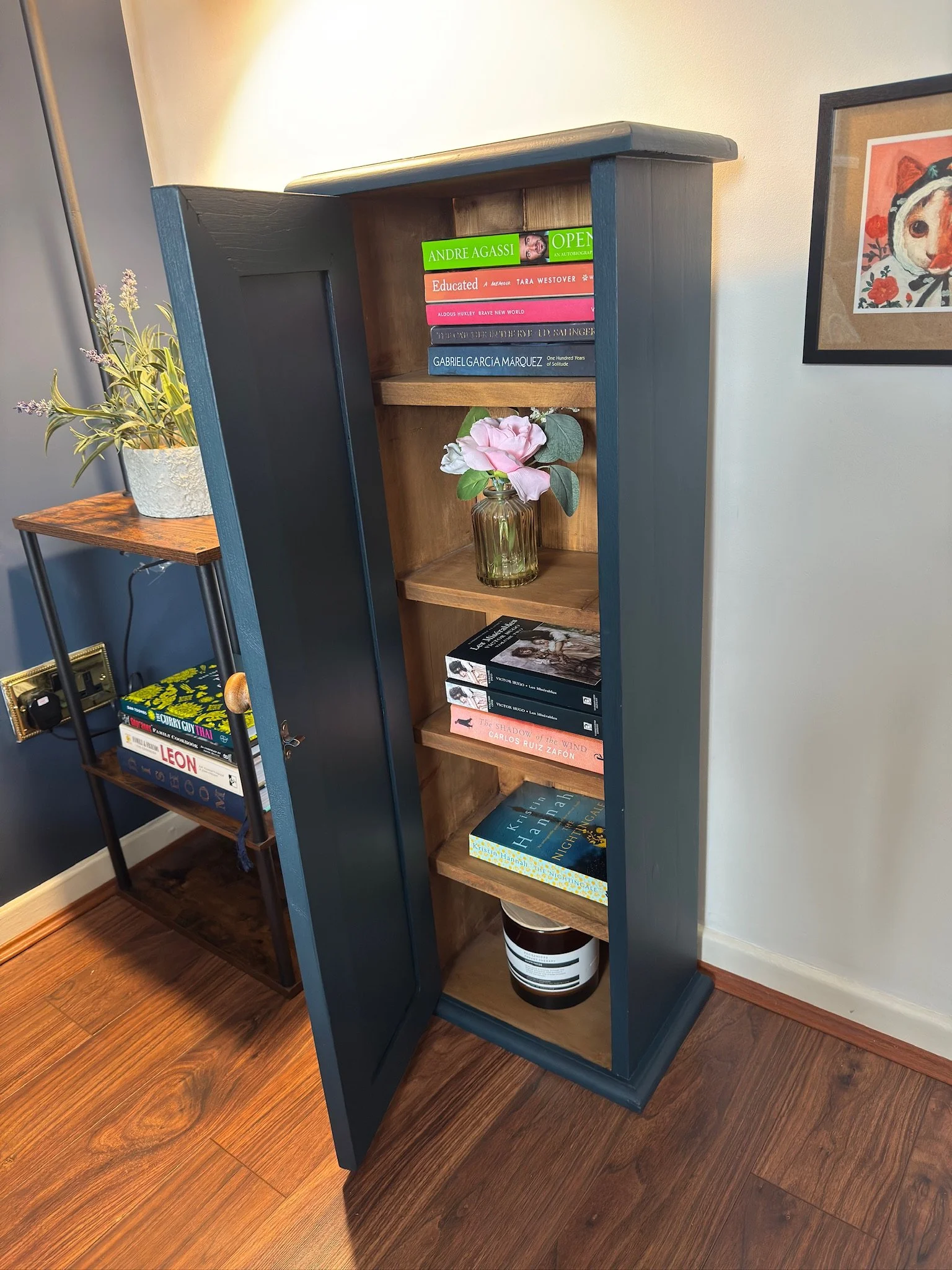 Black and natural wood bookshelf with five shelves, decorated with books, a vase with pink flowers, and a plant on a side table in a cozy room.