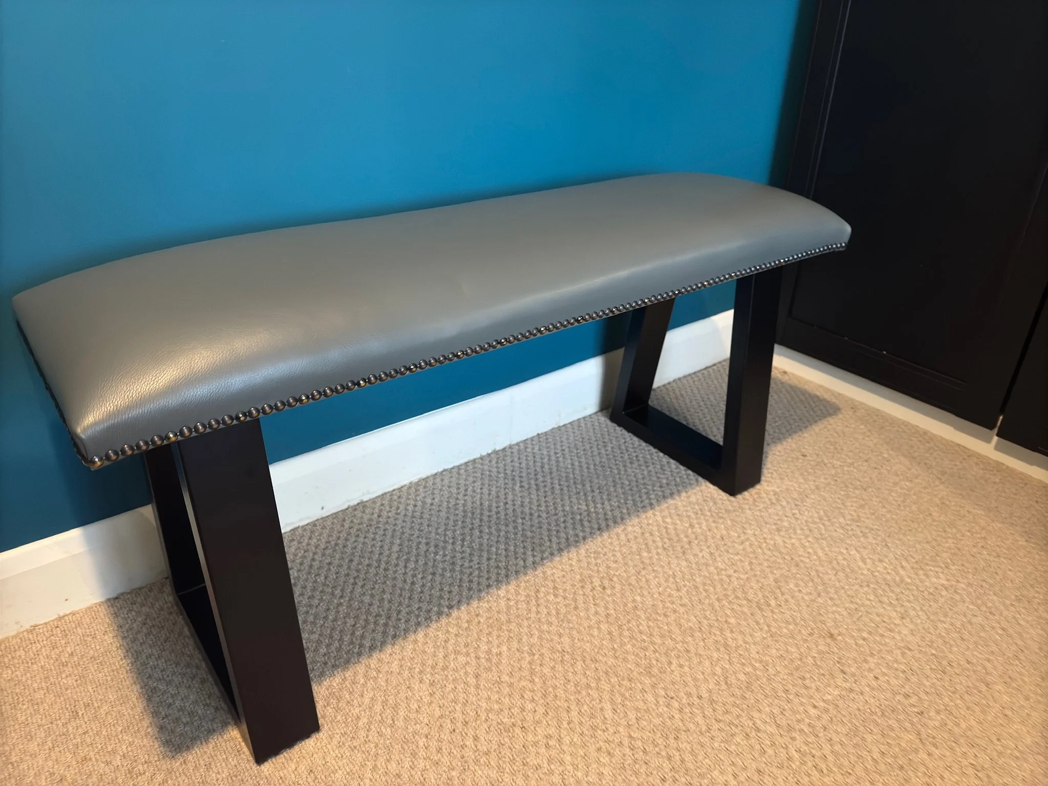 A gray upholstered bench with black wooden legs and metal nailhead trim along the edge, placed against a teal wall near a beige carpeted floor.