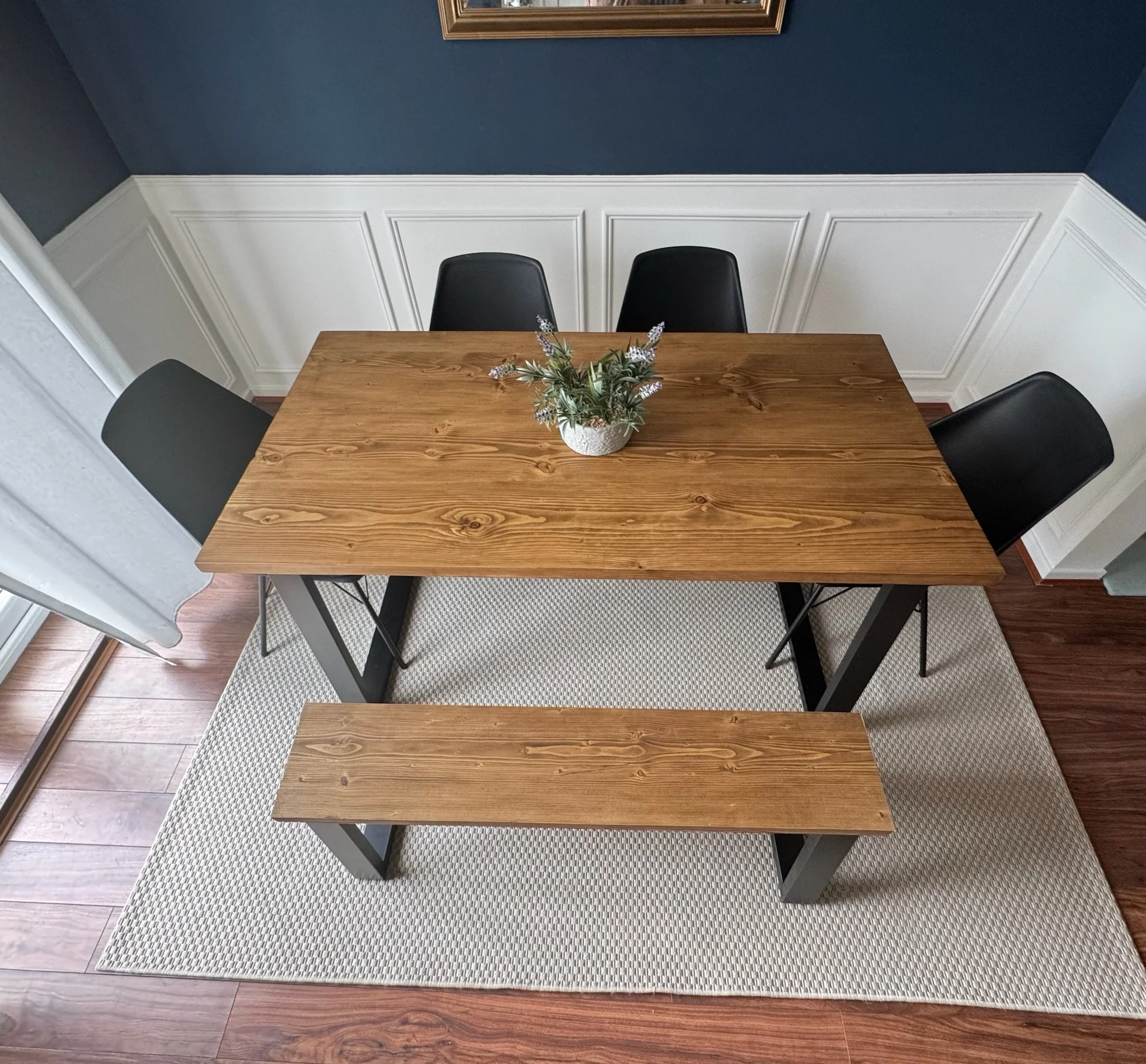 Wood dining table with four black chairs, a small plant in a white pot, and a light-colored rug beneath.