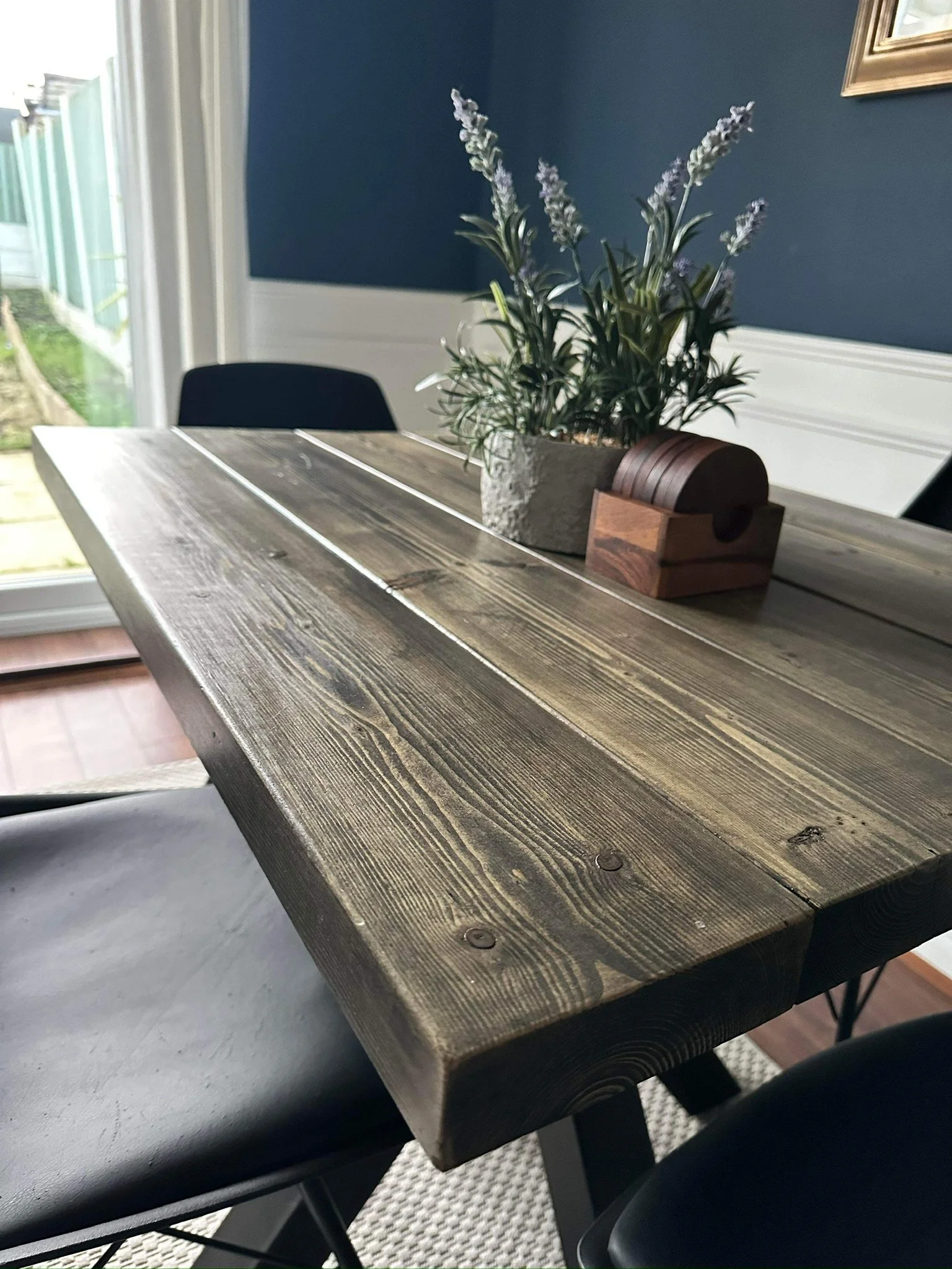 Wooden dining table with a potted lavender plant and a small wooden box on top, with black chairs around it in a dining area near a window.
