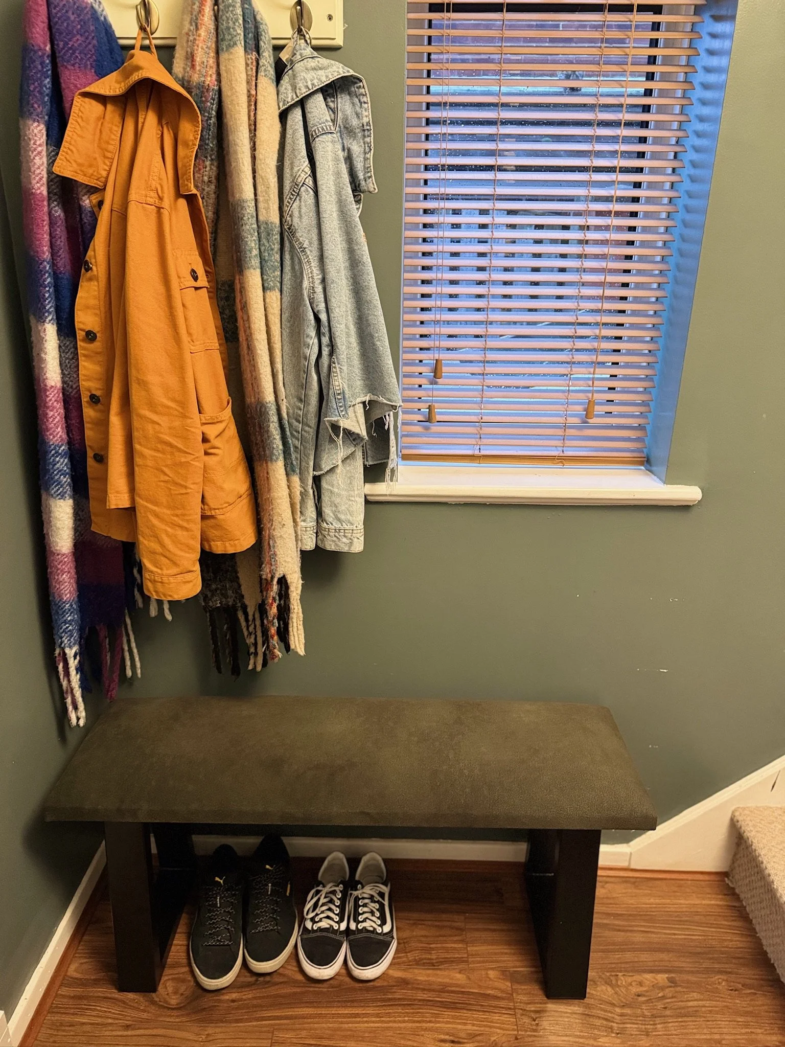 A small entryway with a green wall, a window with wooden blinds, a bench with a brown cushion, and various clothing items hanging on hooks, including a yellow jacket, a plaid blanket, and a denim jacket. Two pairs of sneakers are placed under the ben