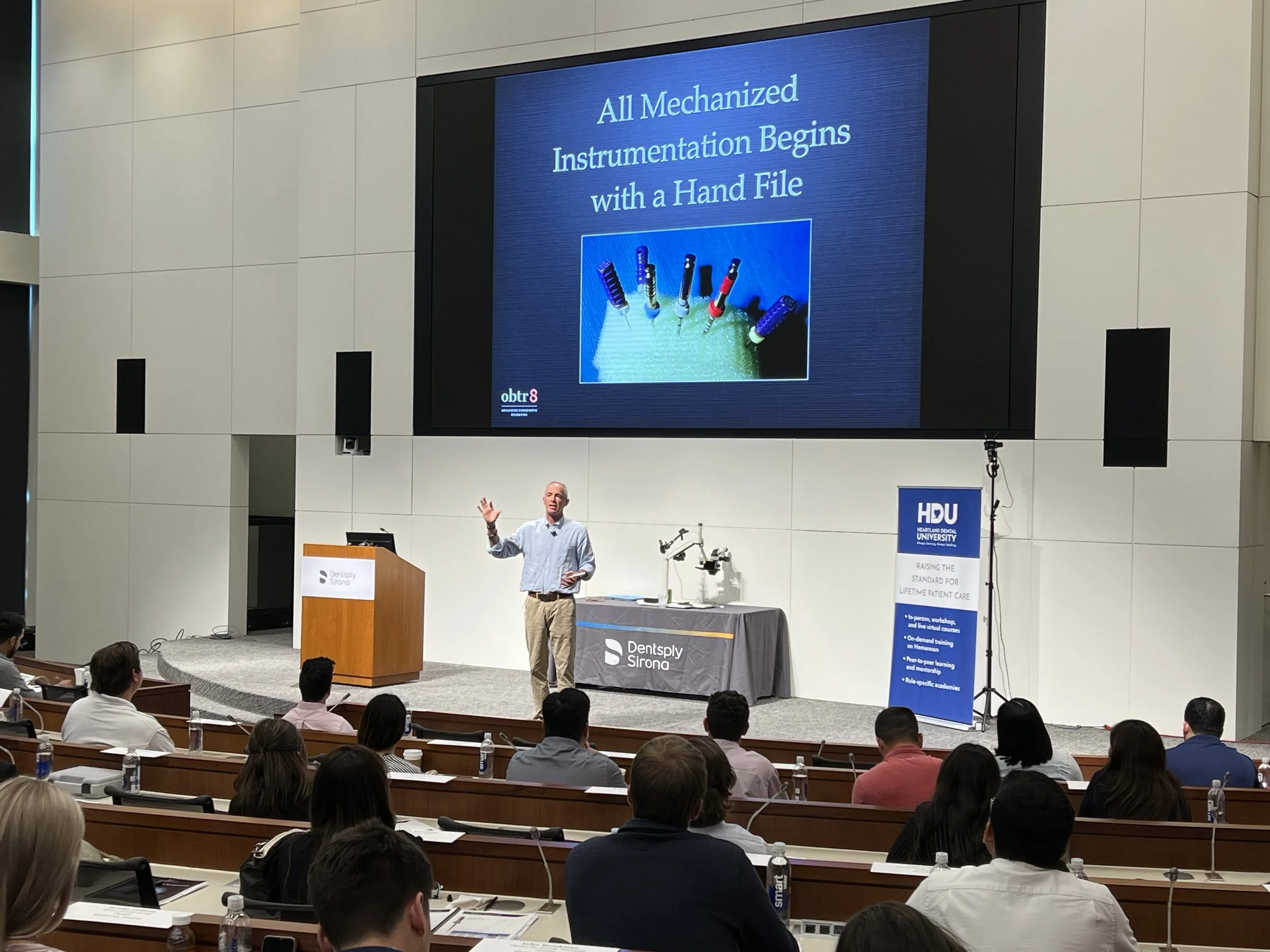 A speaker at a conference presenting on a large screen that reads, 'All mechanized instrumentation begins with a hand file.' The audience is seated, listening, with some taking notes, and the event is hosted at an indoor auditorium.
