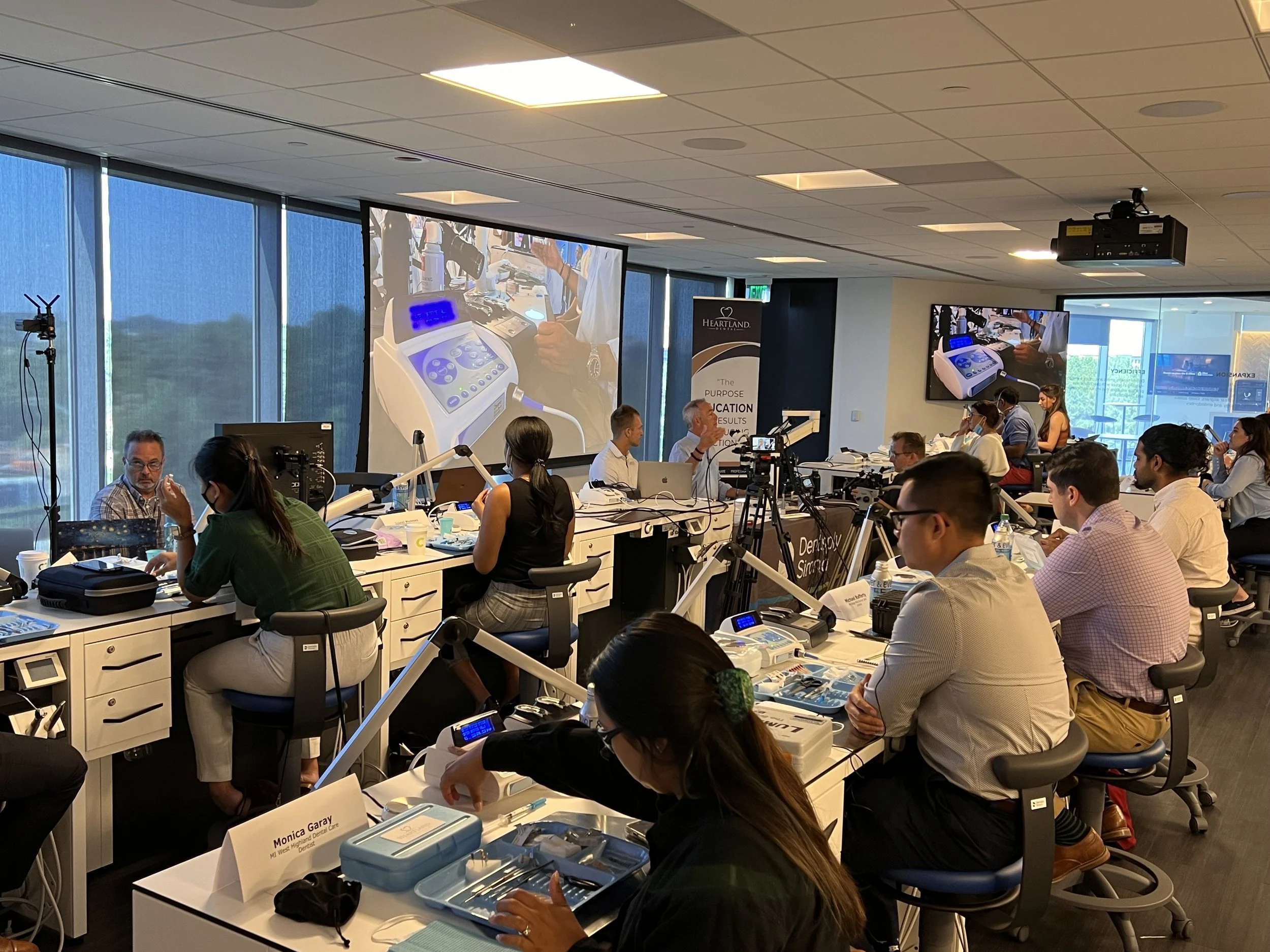 A conference room filled with people attending a workshop or seminar. Several large screens display close-up images of medical or dental equipment. Attendees are seated at tables, some wearing masks, with various supplies and name tags visible. The setting appears professional and educational.