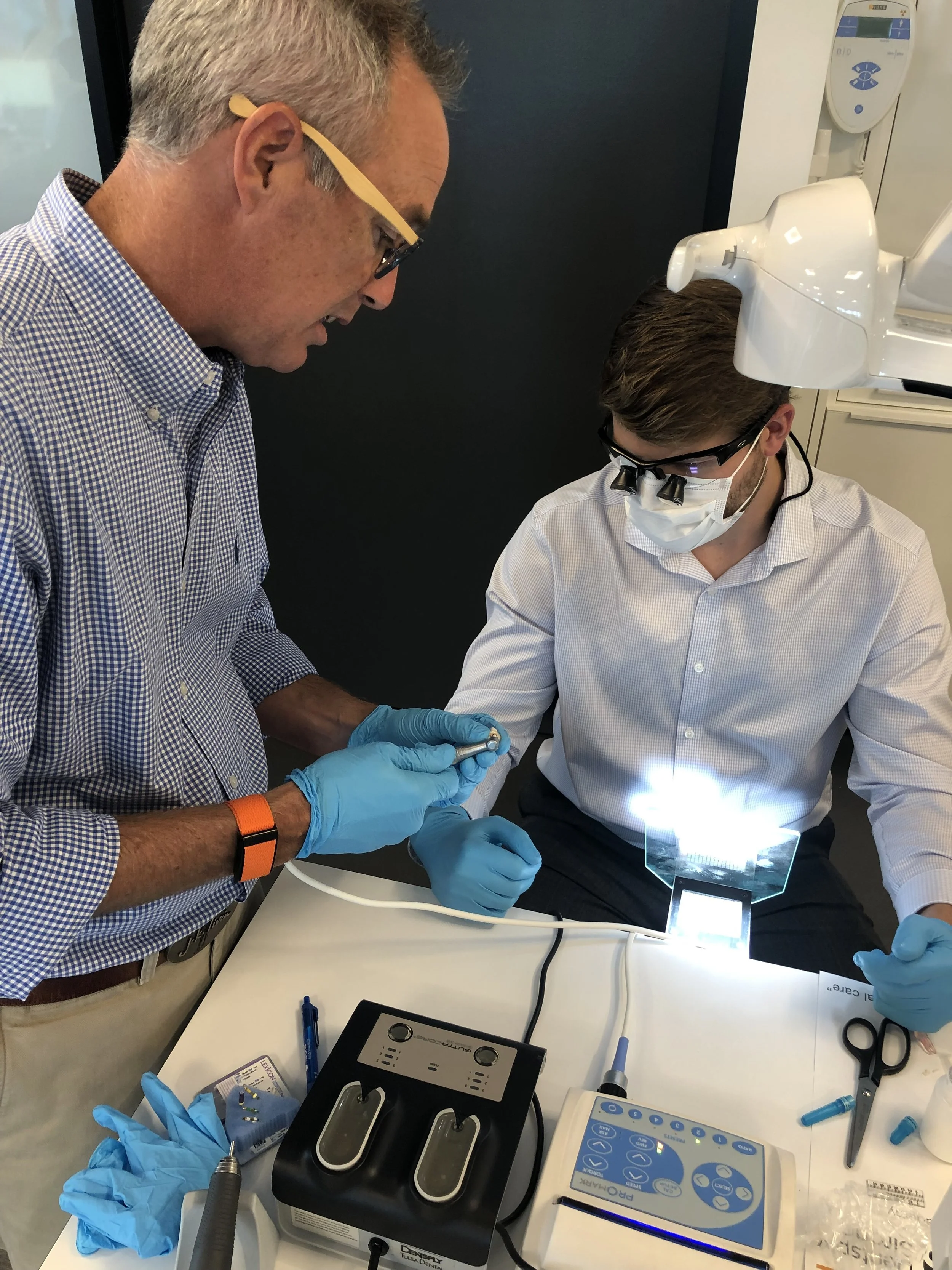 Two scientists wearing protective masks and gloves working with dental equipment in a lab, one holding a small dental tool while the other observes under a magnifying device. Dental tools and equipment are on the table.