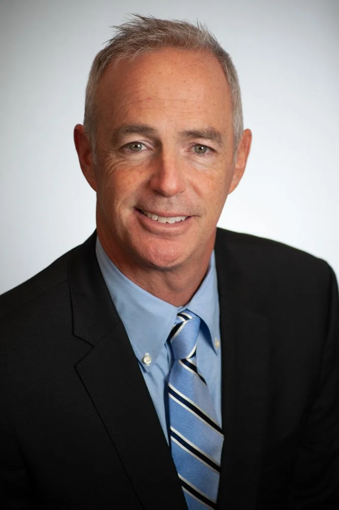 A professional headshot of a middle-aged man with short gray hair, wearing a dark suit, light blue shirt, and a blue striped tie, smiling against a plain light gray background.