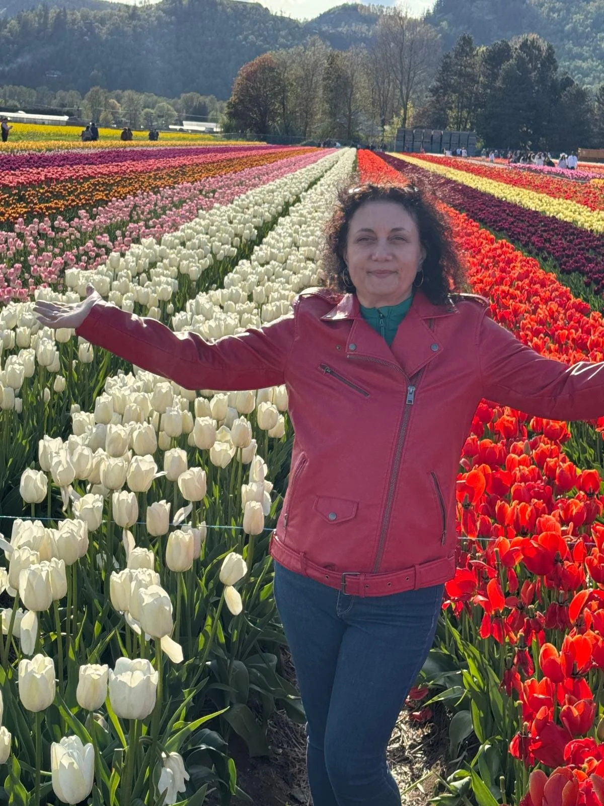 A person in a red jacket posing with arms open in front of a colorful field of tulips, with rows of red, white, pink, and orange flowers. Trees and mountains are visible in the background.