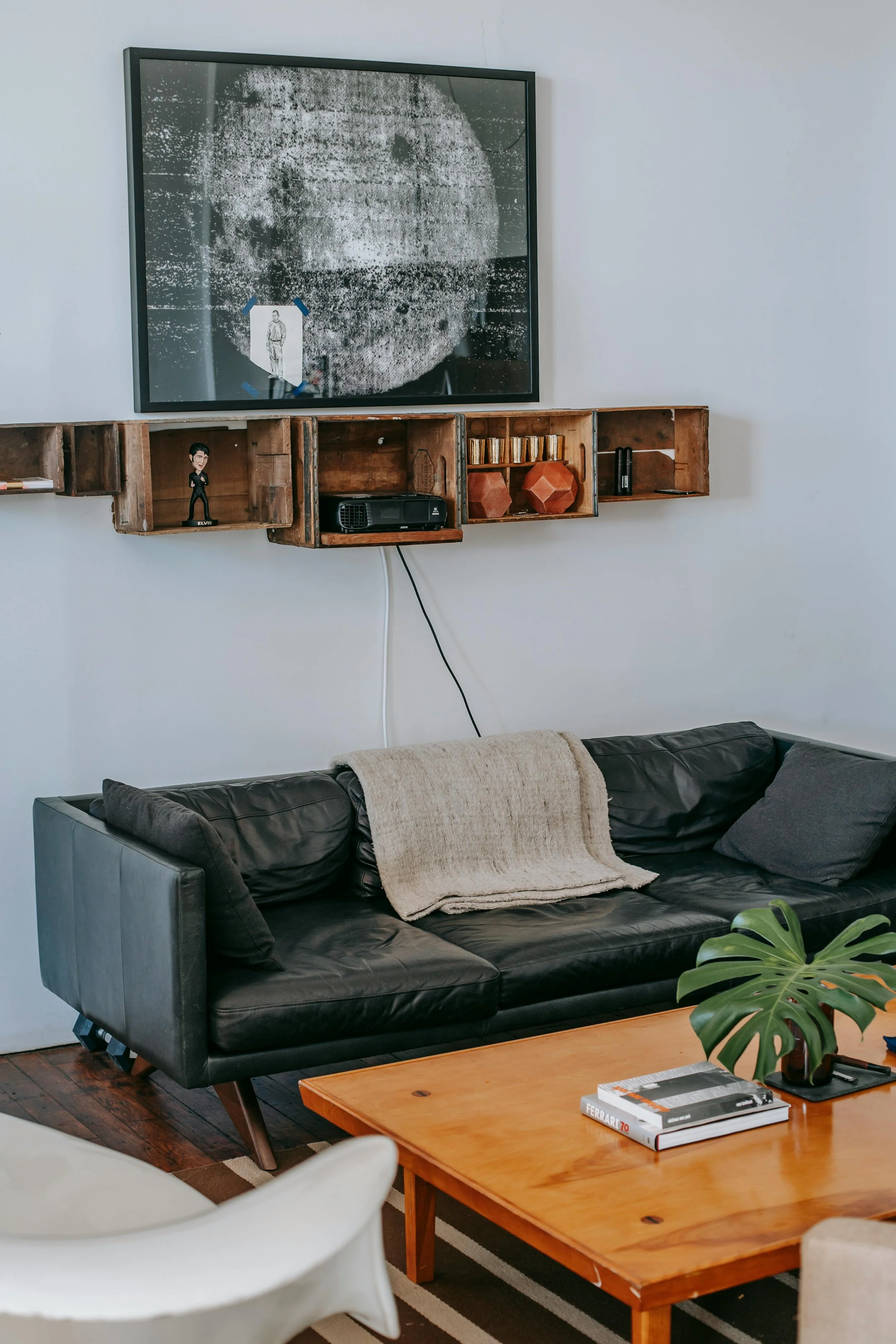Living room with a black leather sofa, wooden crate shelves displaying various items, a framed moon artwork on the wall, and a wooden coffee table with books and a potted plant.