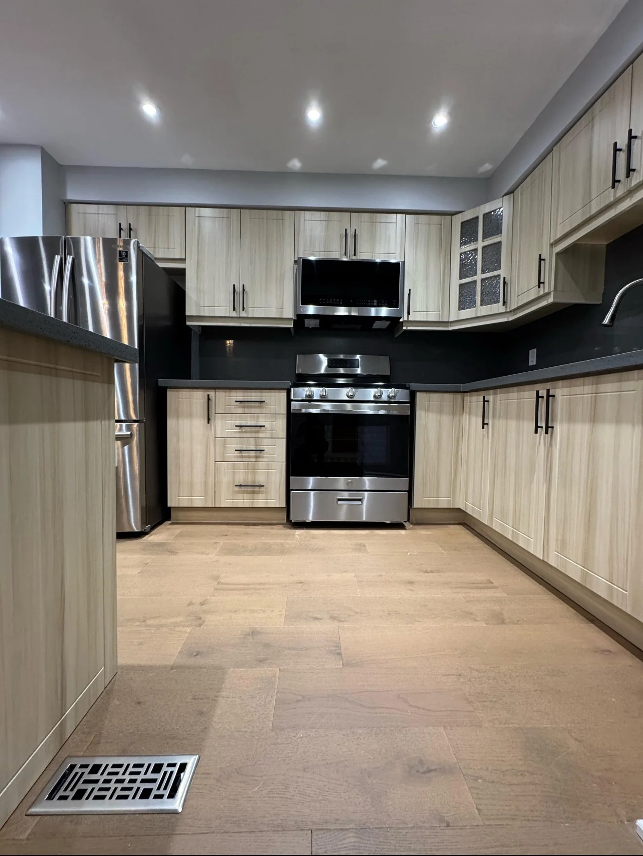 Modern kitchen with stainless steel appliances, light wood cabinets, and wood flooring. Features a refrigerator, over-the-range microwave, oven, and under-cabinet lighting.