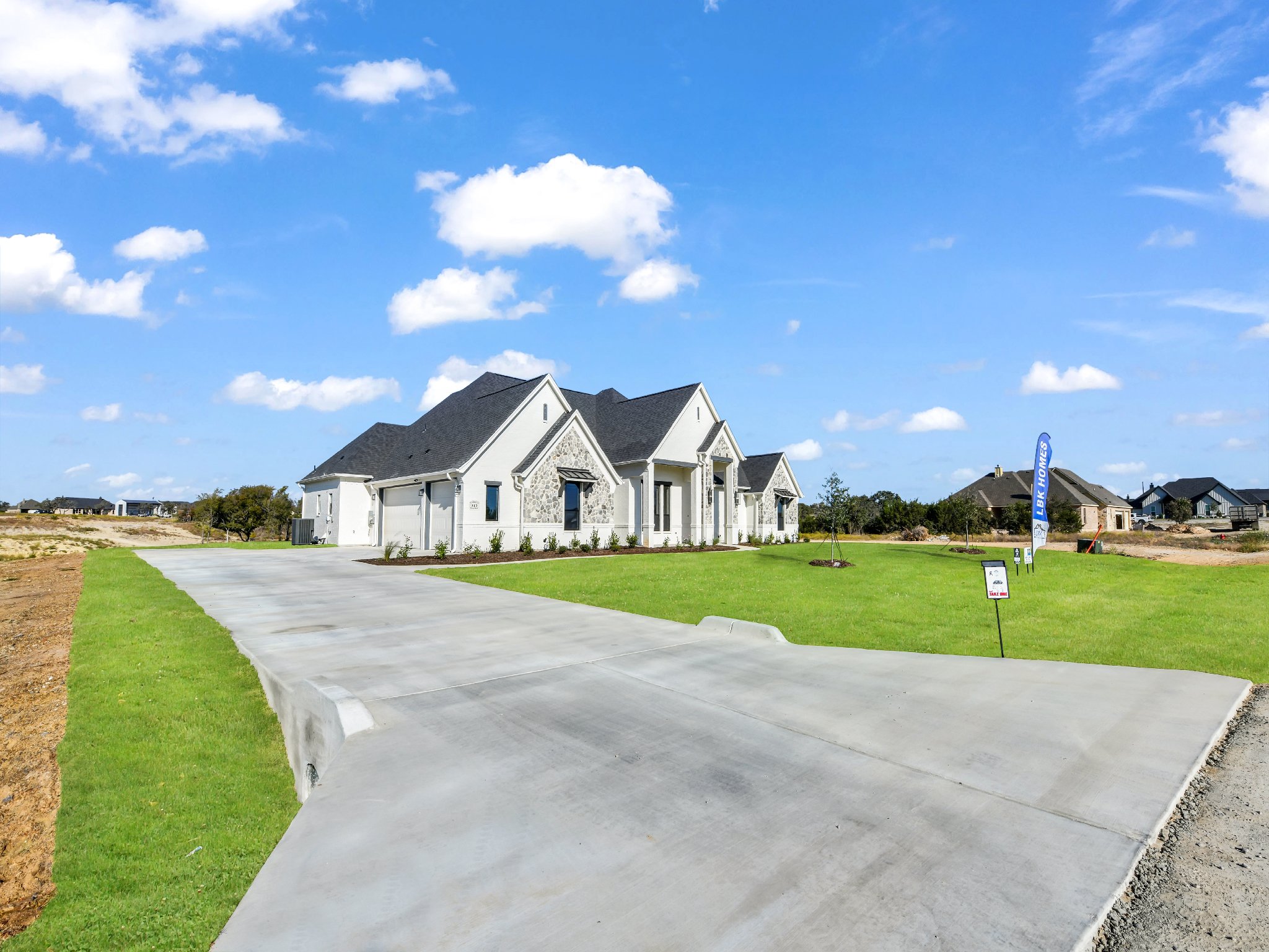Newly constructed white house with black roof, expansive driveway, green lawn, and a 'for sale' sign in front, under a partly cloudy blue sky.