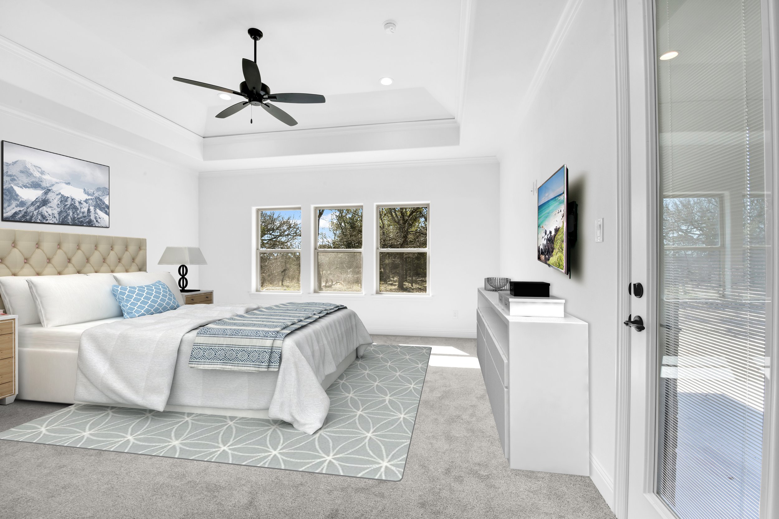 Modern bedroom with a large bed, white bedding, patterned pillows, a nightstand with a lamp, three windows showing trees outside, a wall-mounted TV, and sliding glass door with blinds.