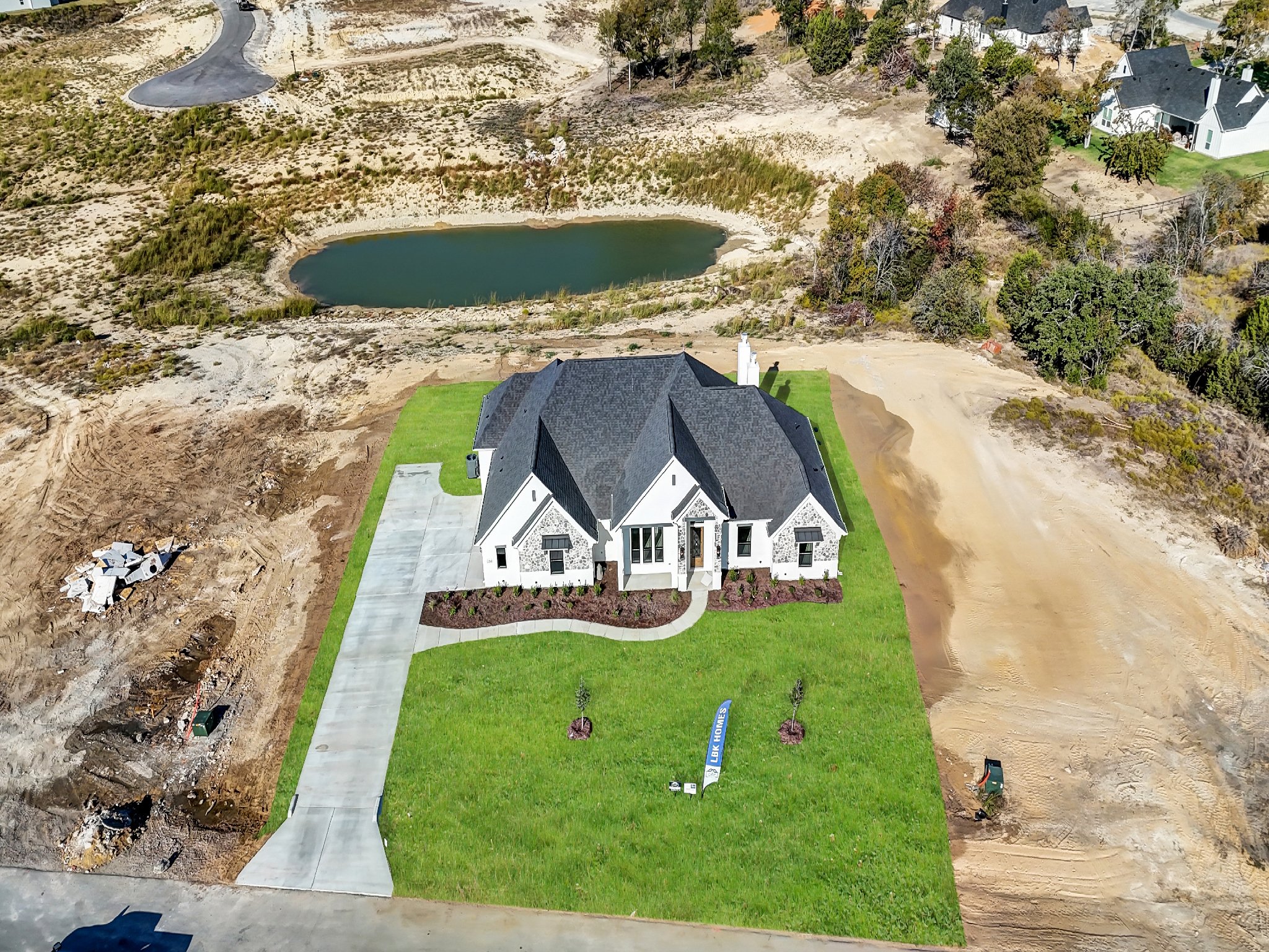 A newly built house with a gray roof and white exterior, surrounded by a green lawn, under construction with dirt and construction materials nearby, near a small pond in a semi-rural area.