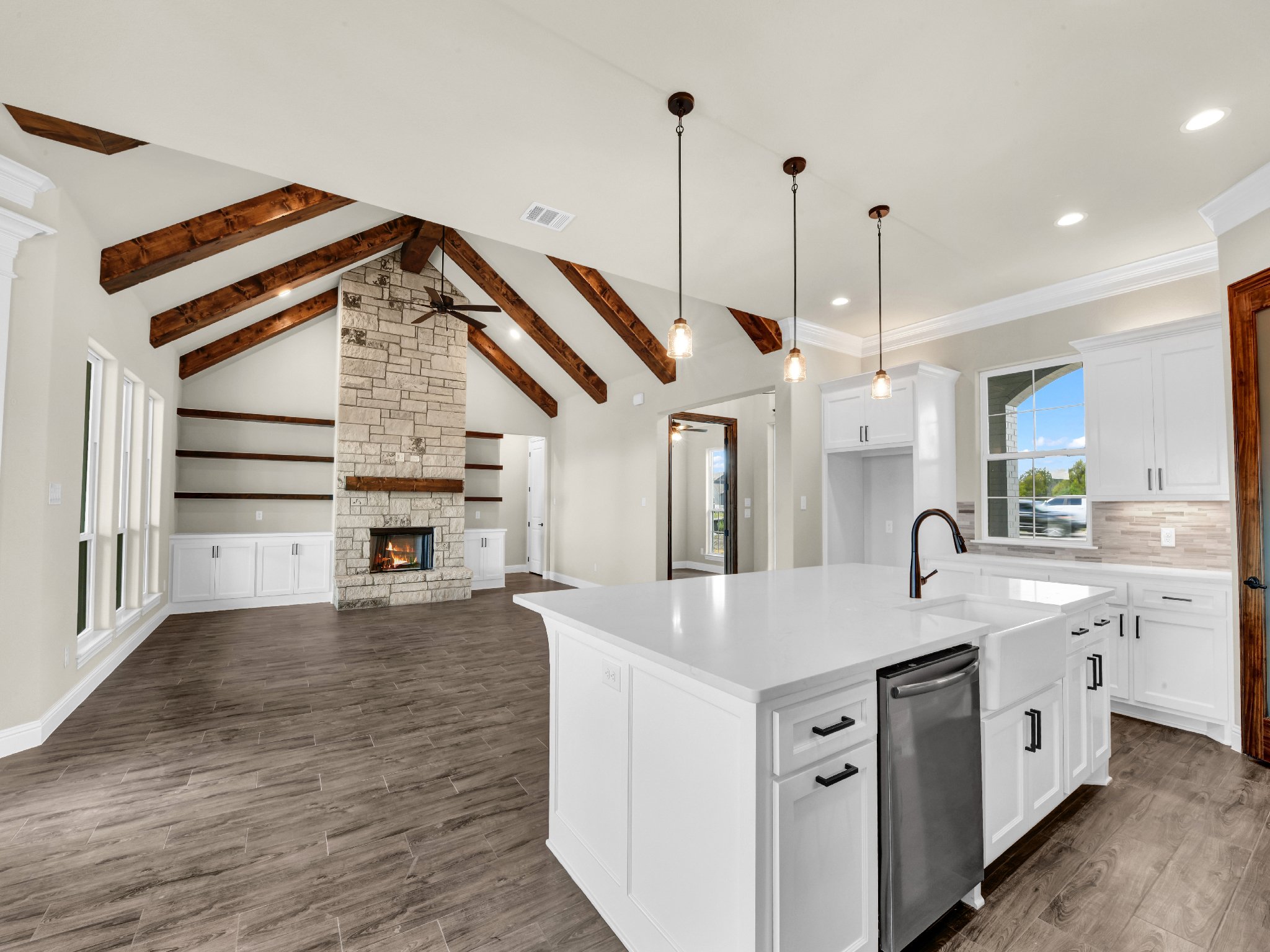 Open-concept living room and kitchen with vaulted ceiling, exposed wooden beams, stone fireplace, white cabinets, and island with black hardware, hardwood flooring, and large windows.